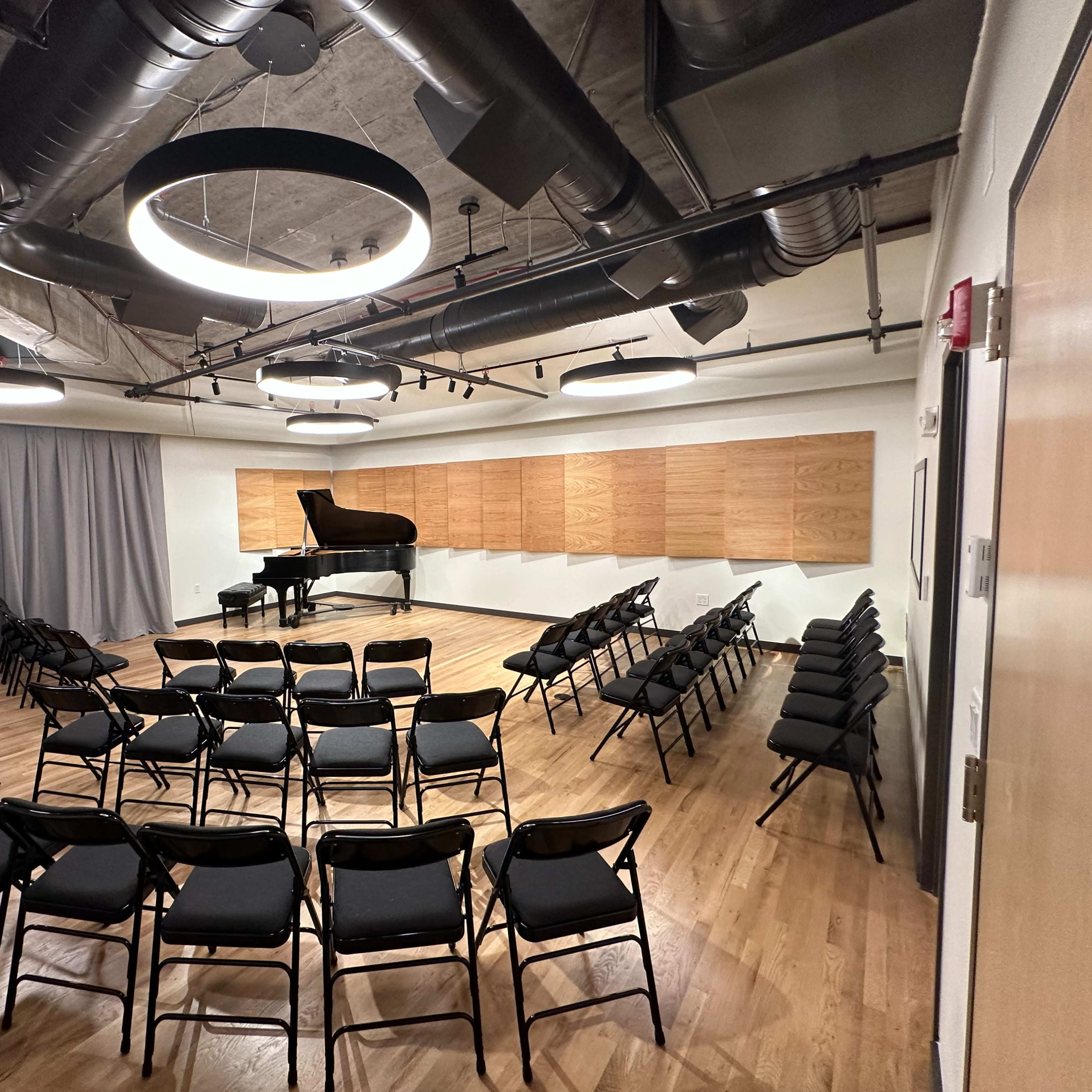 A small performance space features a grand piano, surrounded by neatly arranged black chairs and wooden paneling on the wall.