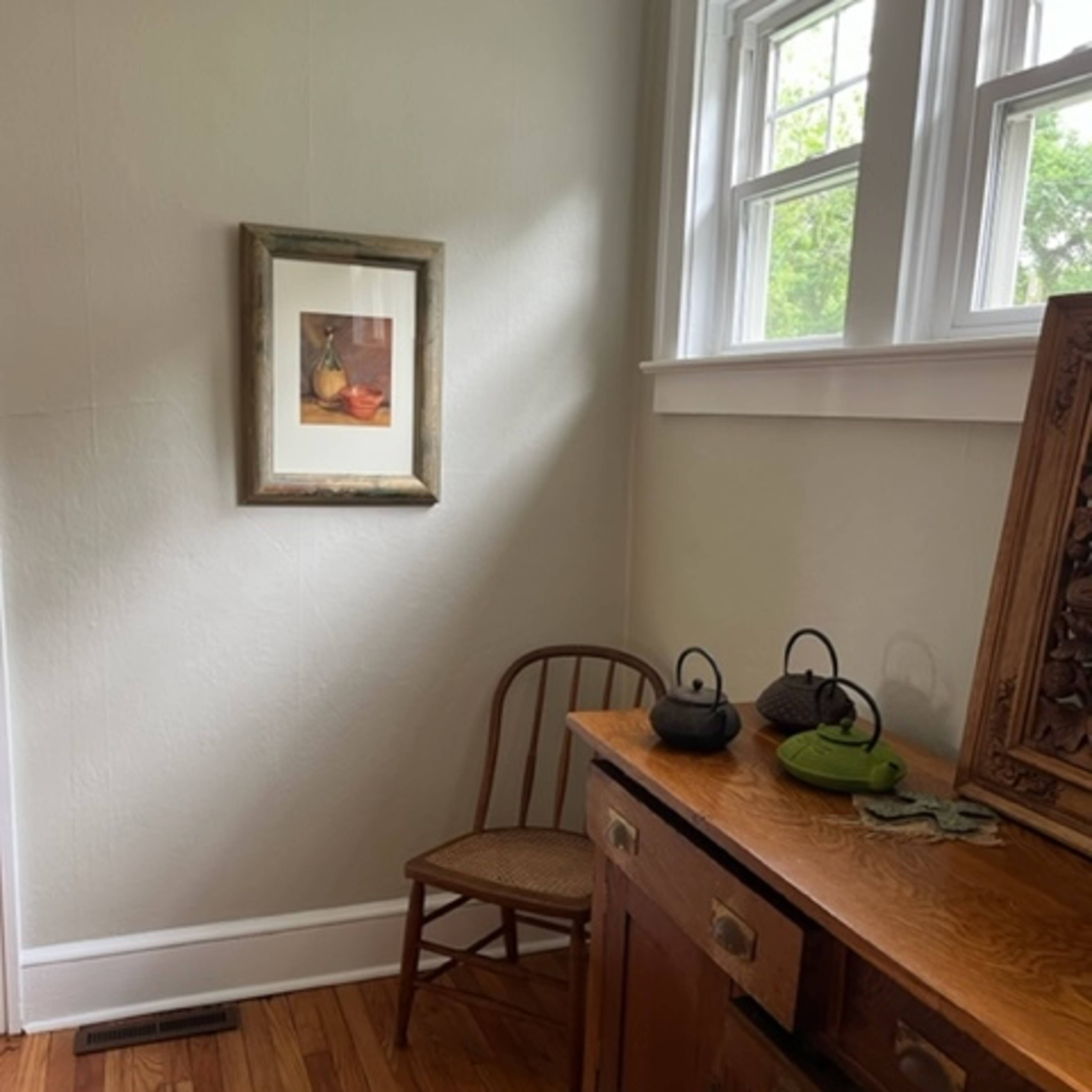 A corner of a room with a chair, a wooden sideboard, a framed picture on the wall, and two teapots on the sideboard.