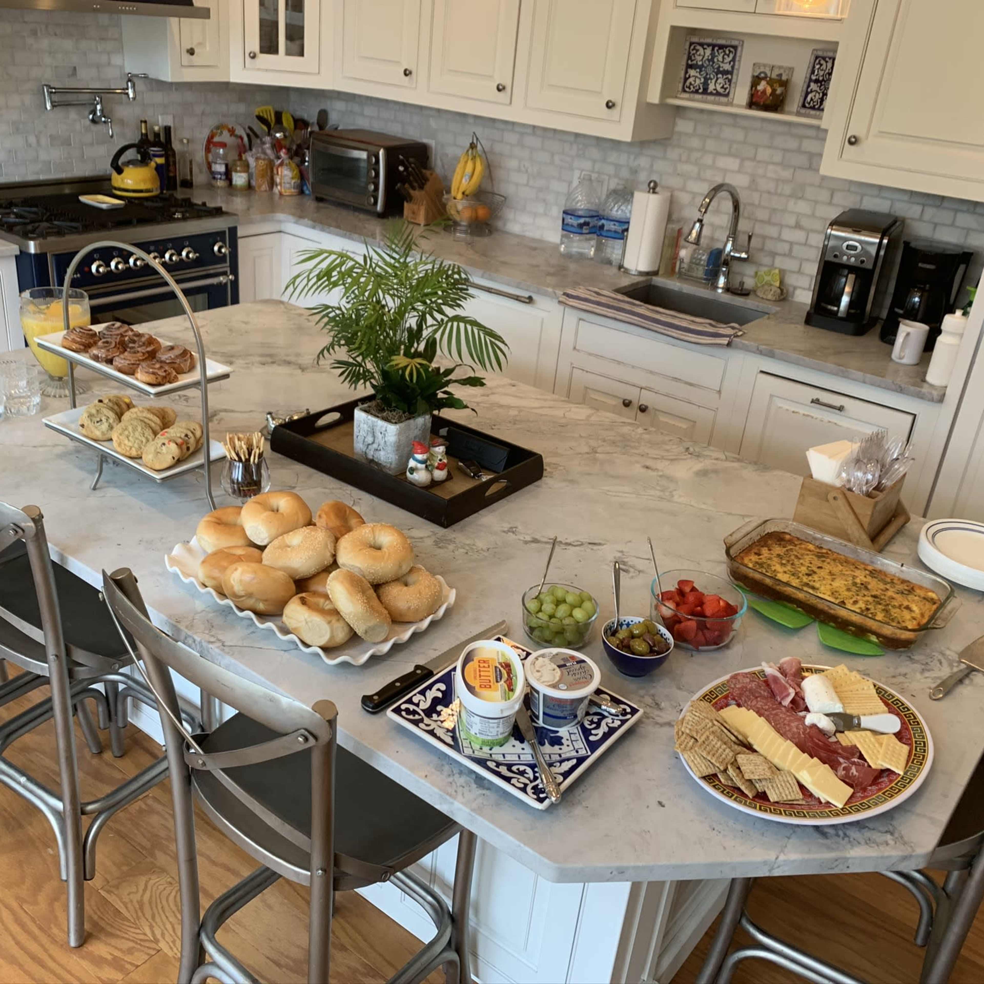 A spacious kitchen island is laden with an assortment of bagels, pastries, cheeses, olives, and fresh fruit, surrounded by several dining chairs.