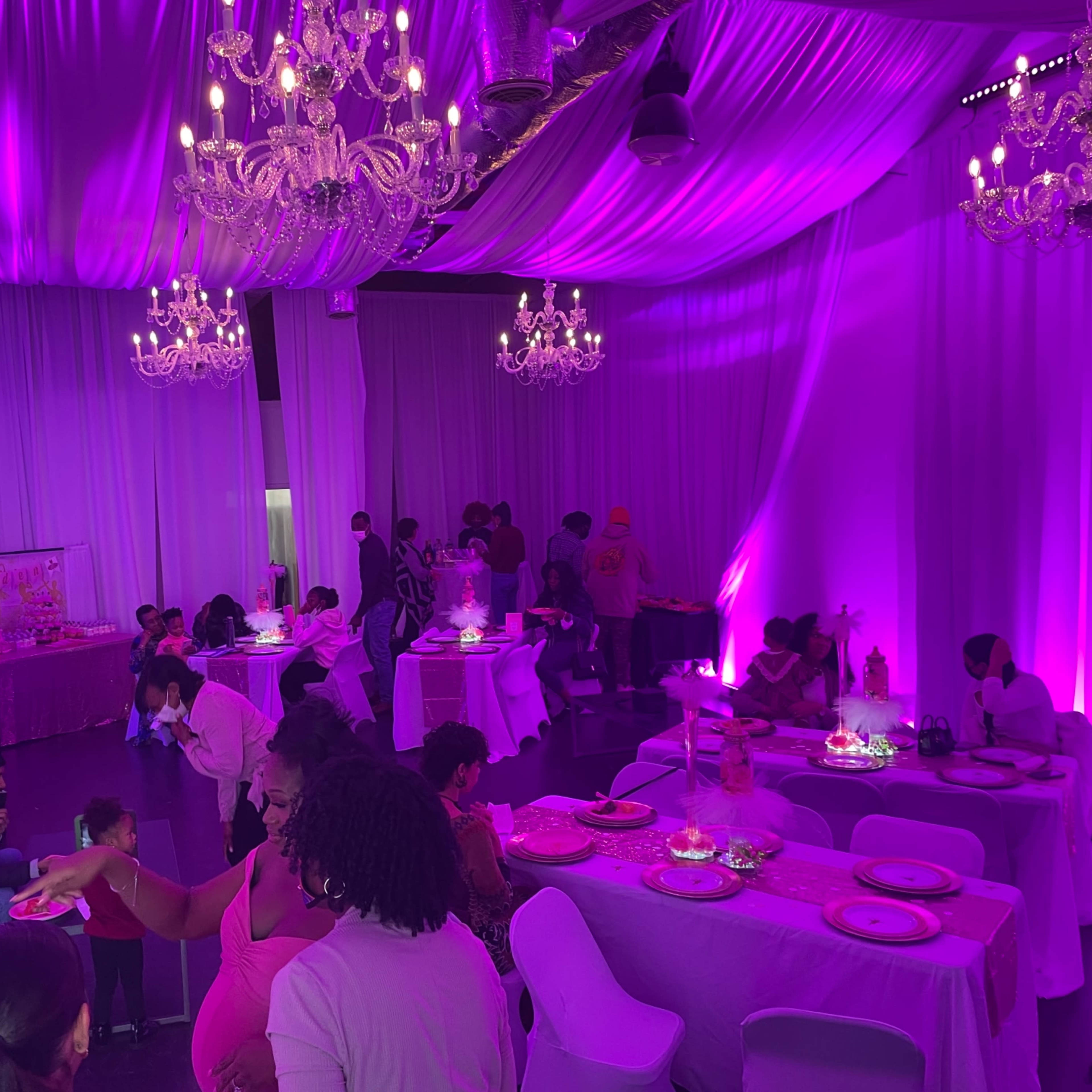 An elegantly decorated indoor event space with purple lighting, chandeliers, and guests mingling around tables set for dining.