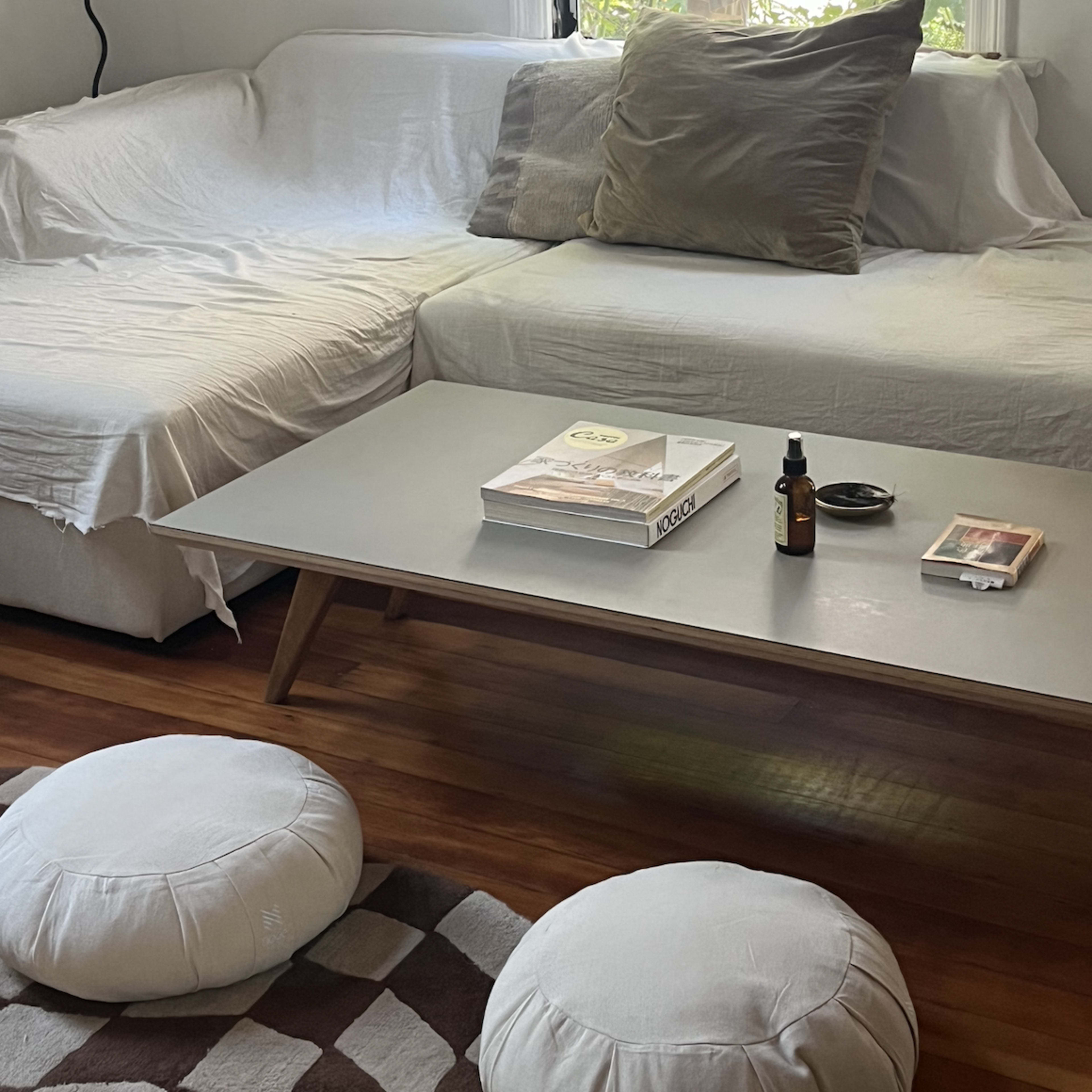 A living room features a white sofa with pillows, a light wooden coffee table displaying a magazine and a small bottle, and two round cushions on a patterned rug.