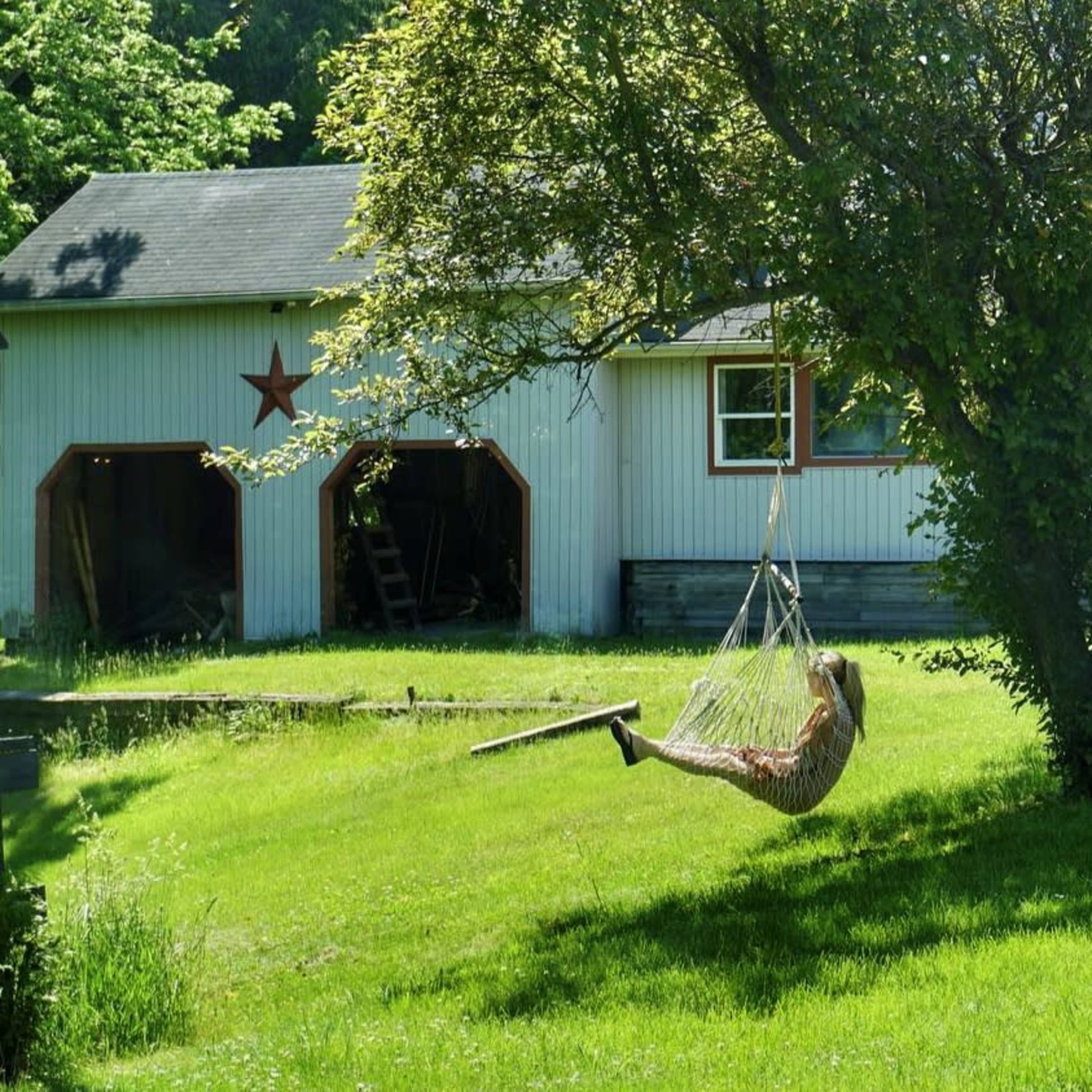 A person swings gently in a hammock under a tree beside a barn with a star decoration on its side.