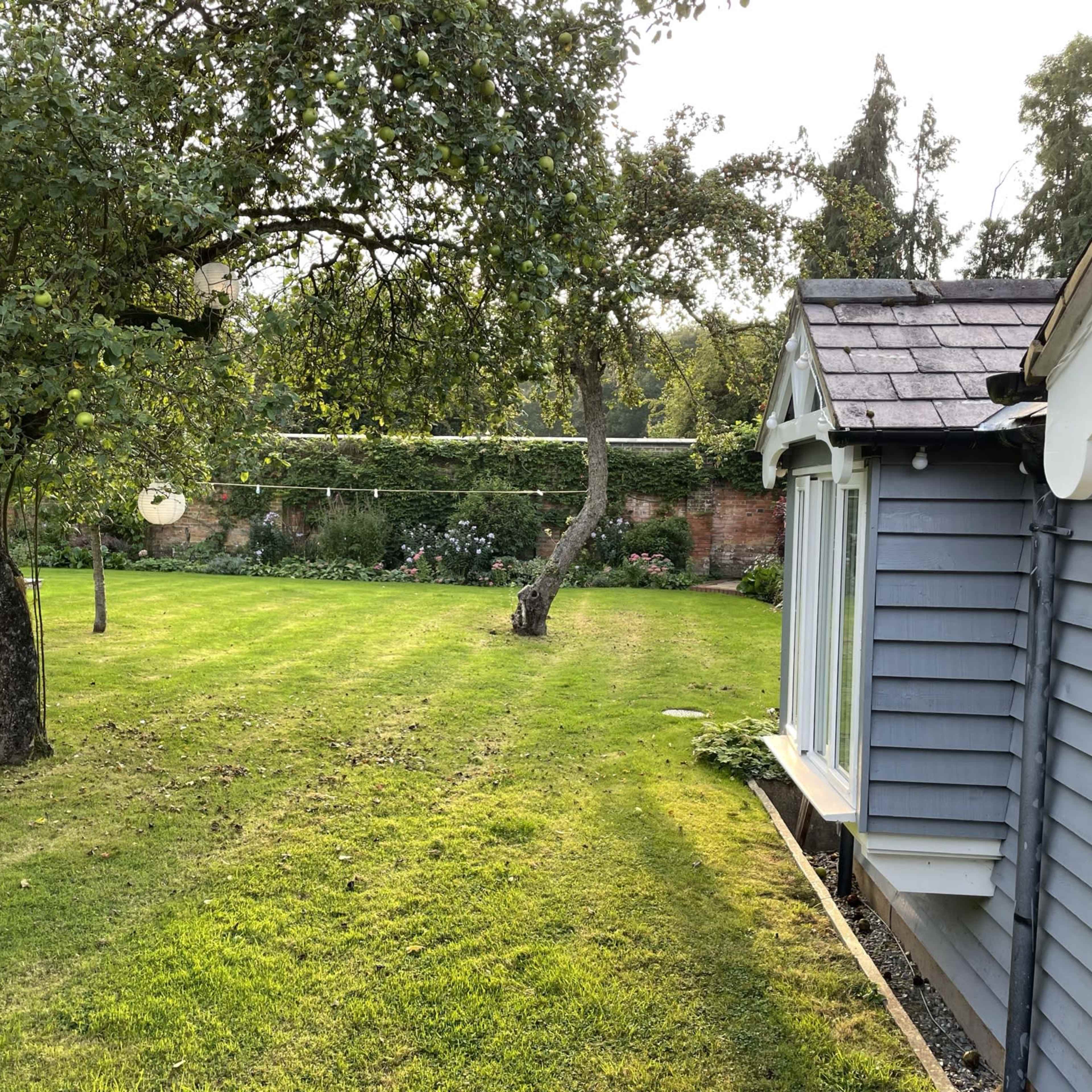 A well-maintained lawn with a gray wooden house, bordered by trees and a garden wall in the background.