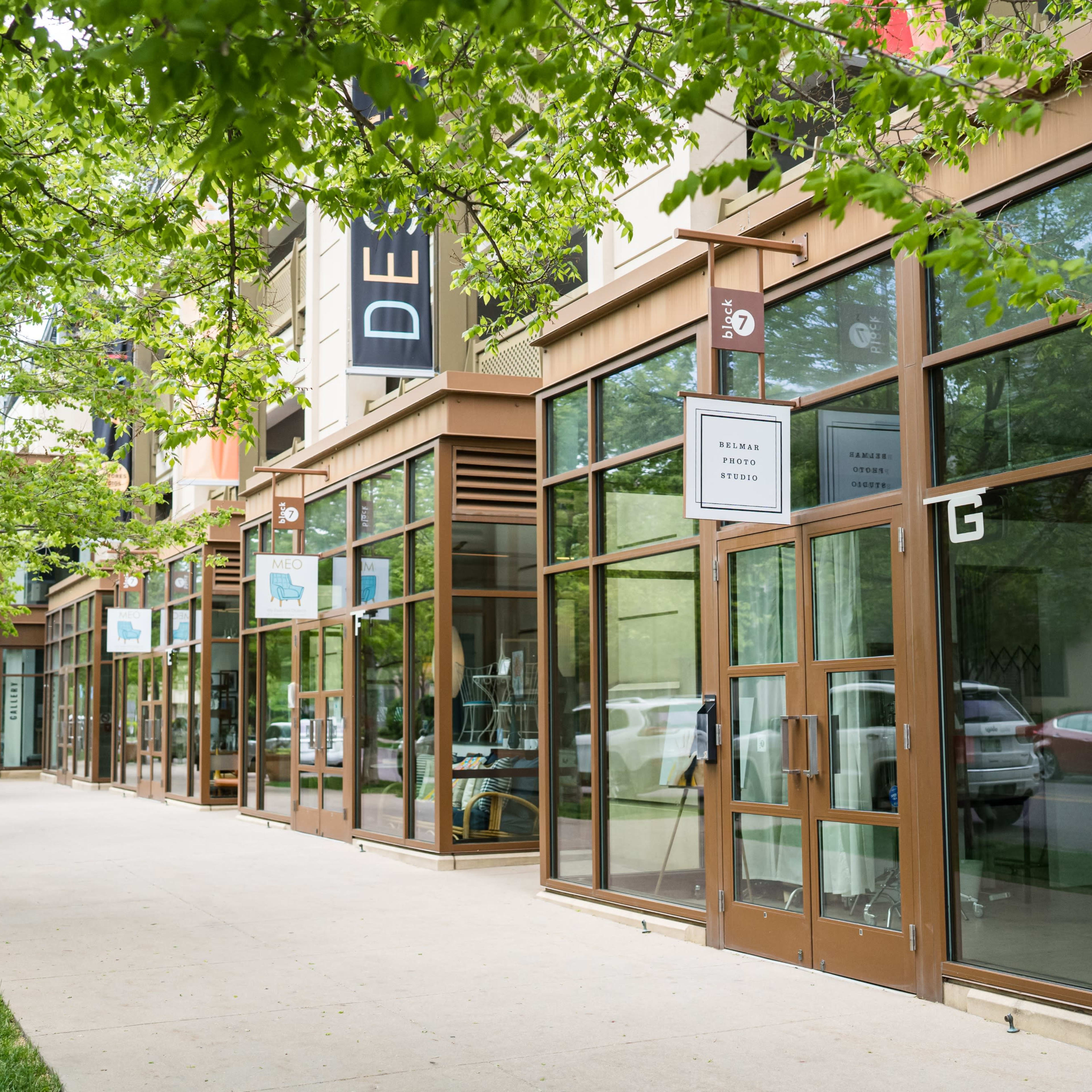A row of storefronts with large windows and greenery along the sidewalk.
