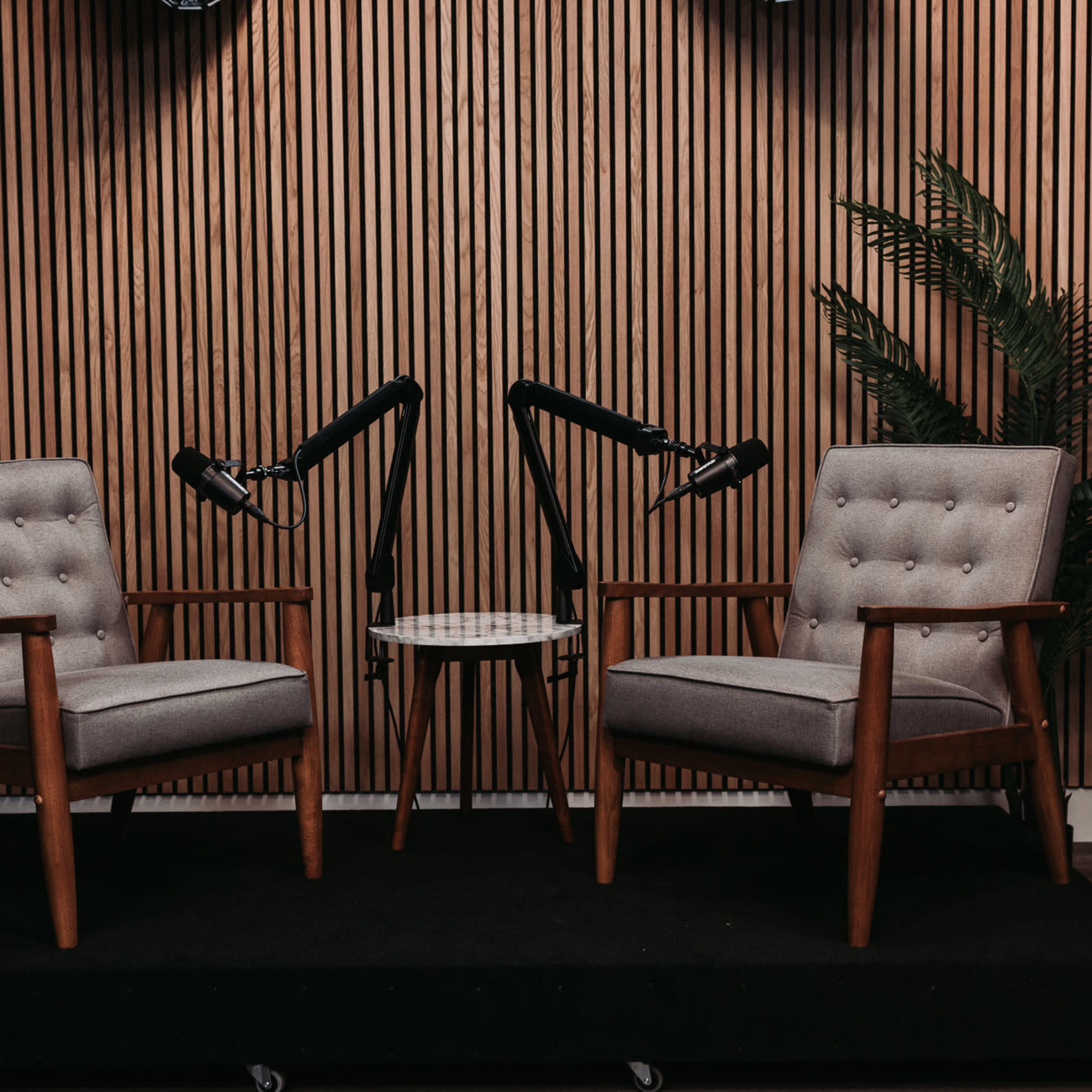 Two upholstered chairs with wooden frames facing each other, accompanied by a small round table and a potted plant, set against a backdrop of vertical wooden slats.