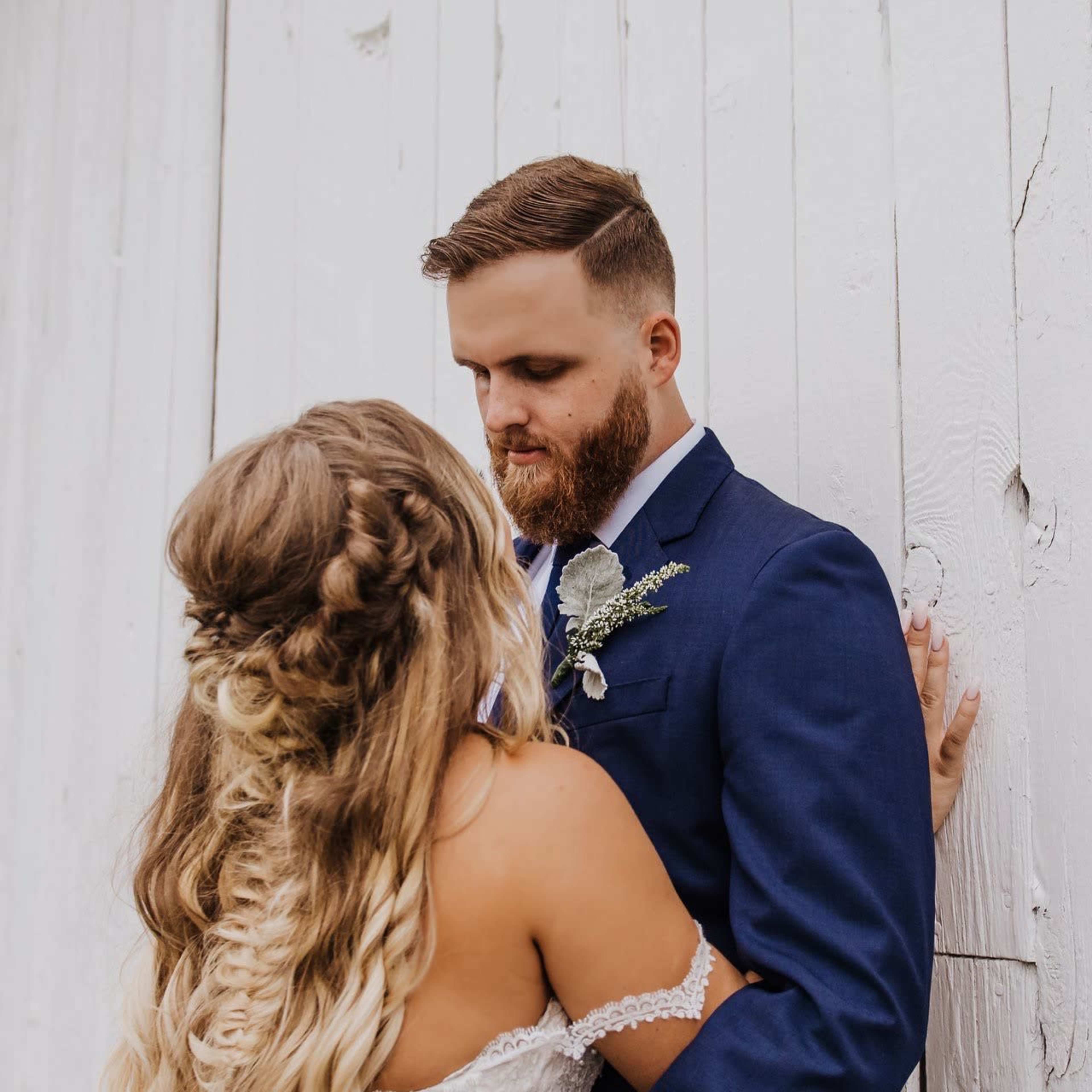 A couple stands closely together, with the woman in a wedding dress and the man in a blue suit, leaning against a white wooden wall.