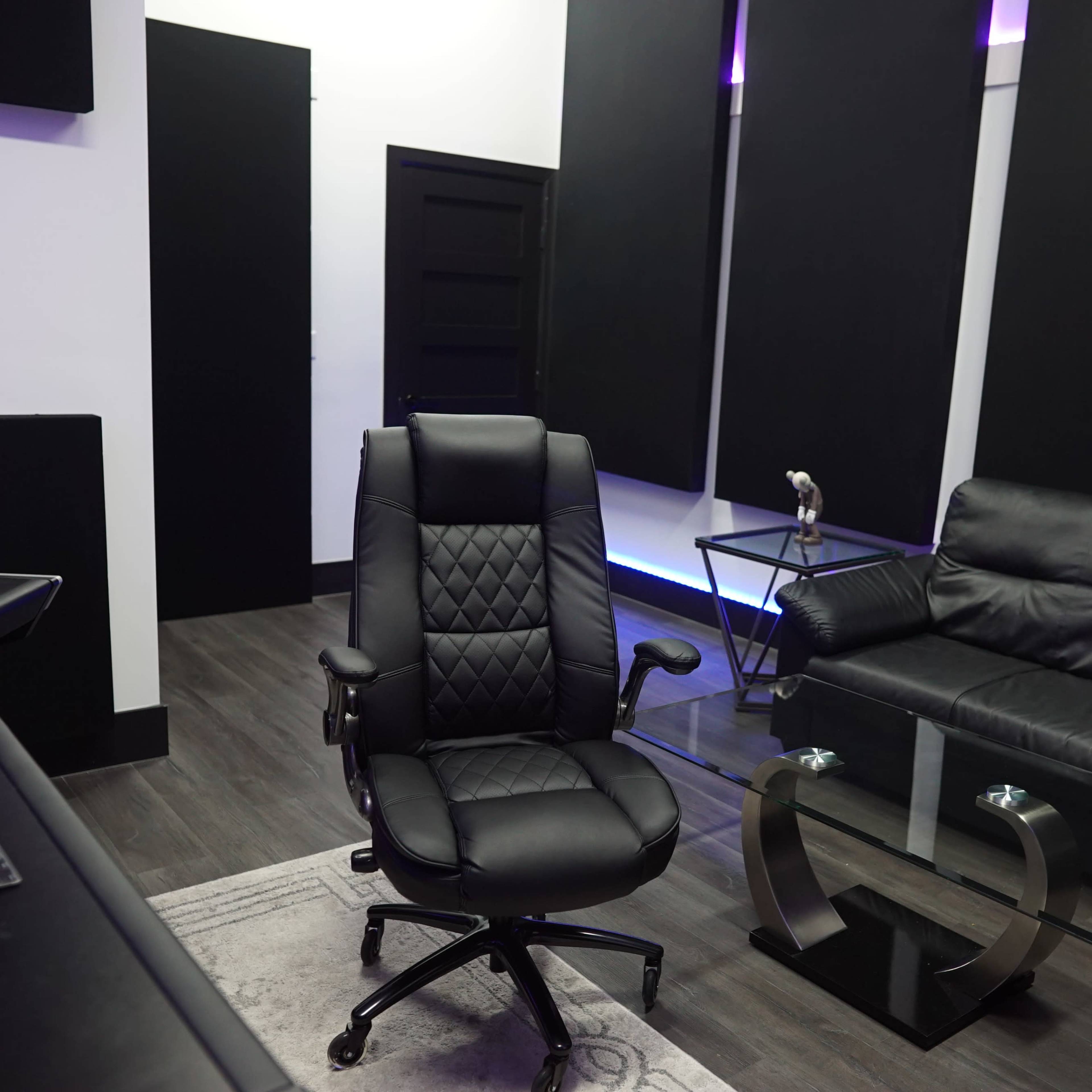 A modern studio room featuring a black ergonomic office chair, a sleek black couch, and a glass coffee table against a backdrop of dark walls and ambient lighting.