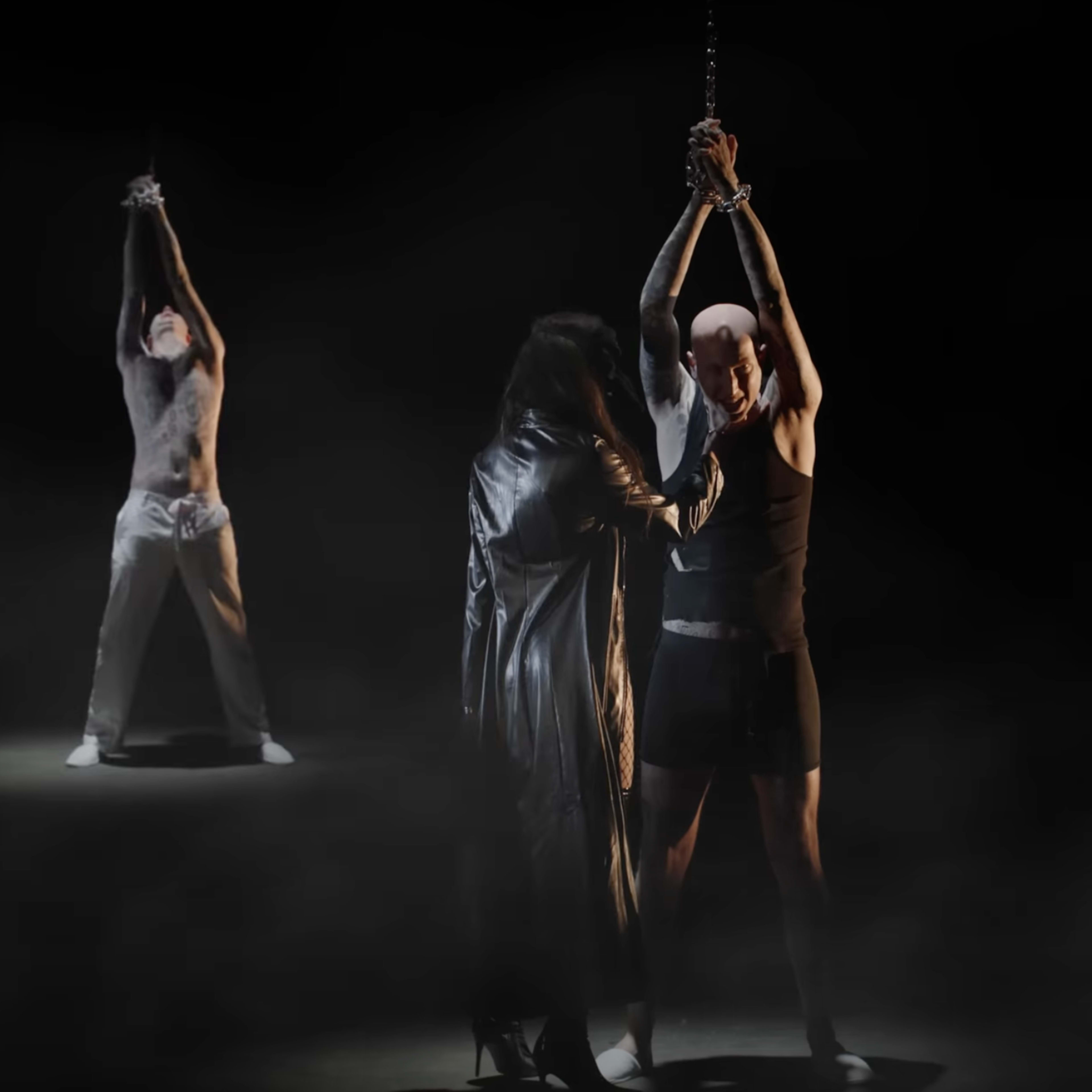 Two figures in a dark setting, one holding the other's wrists above their head, with several shadowy figures in the background.