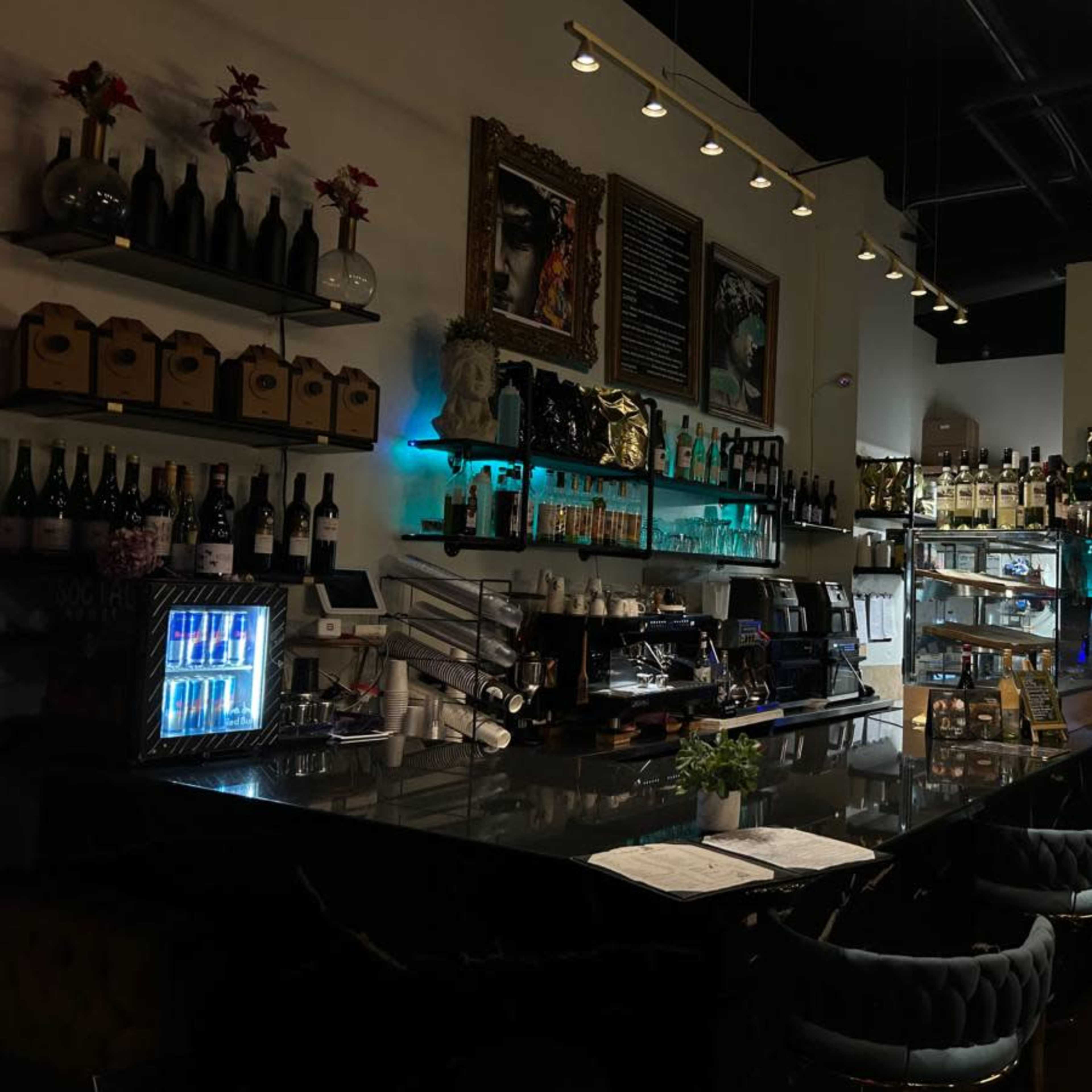 A dimly lit bar counter with shelves displaying various bottles and a coffee machine, accompanied by a menu board and decorative art on the walls.