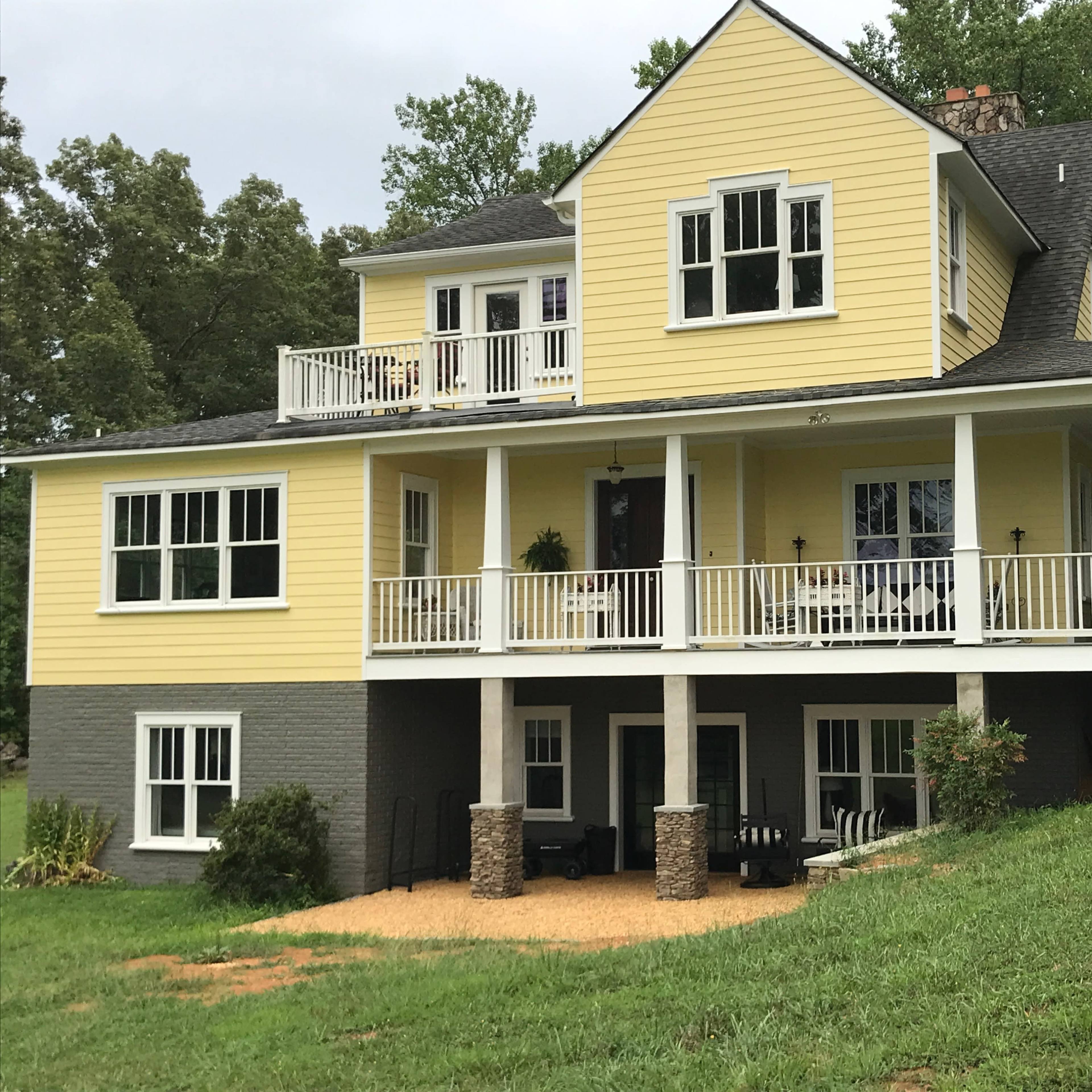 A two-story yellow house with a grey stone foundation and a front porch is situated on a grassy slope surrounded by trees.