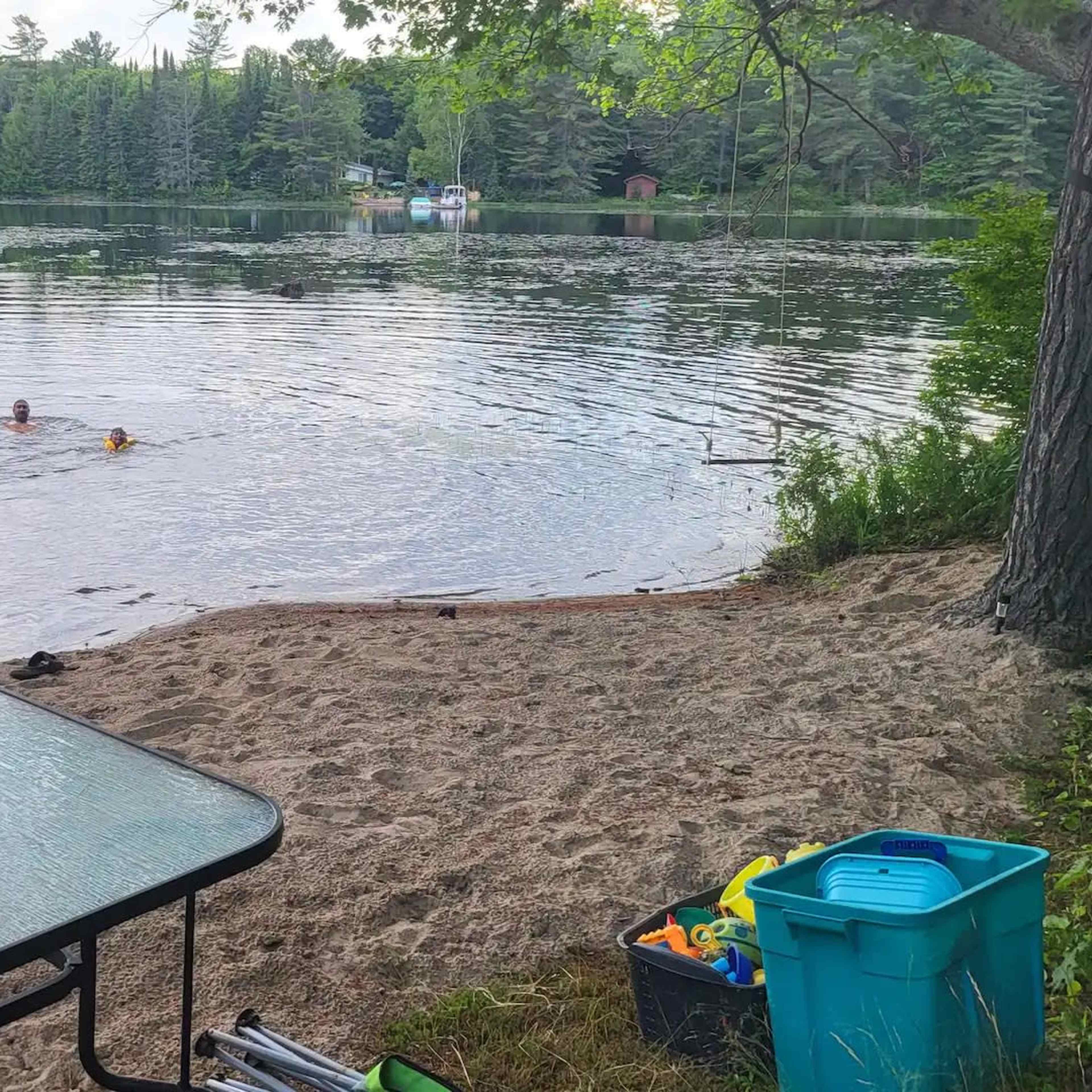 A sandy area near a calm lake features a table and a blue storage bin, while two people swim in the water.