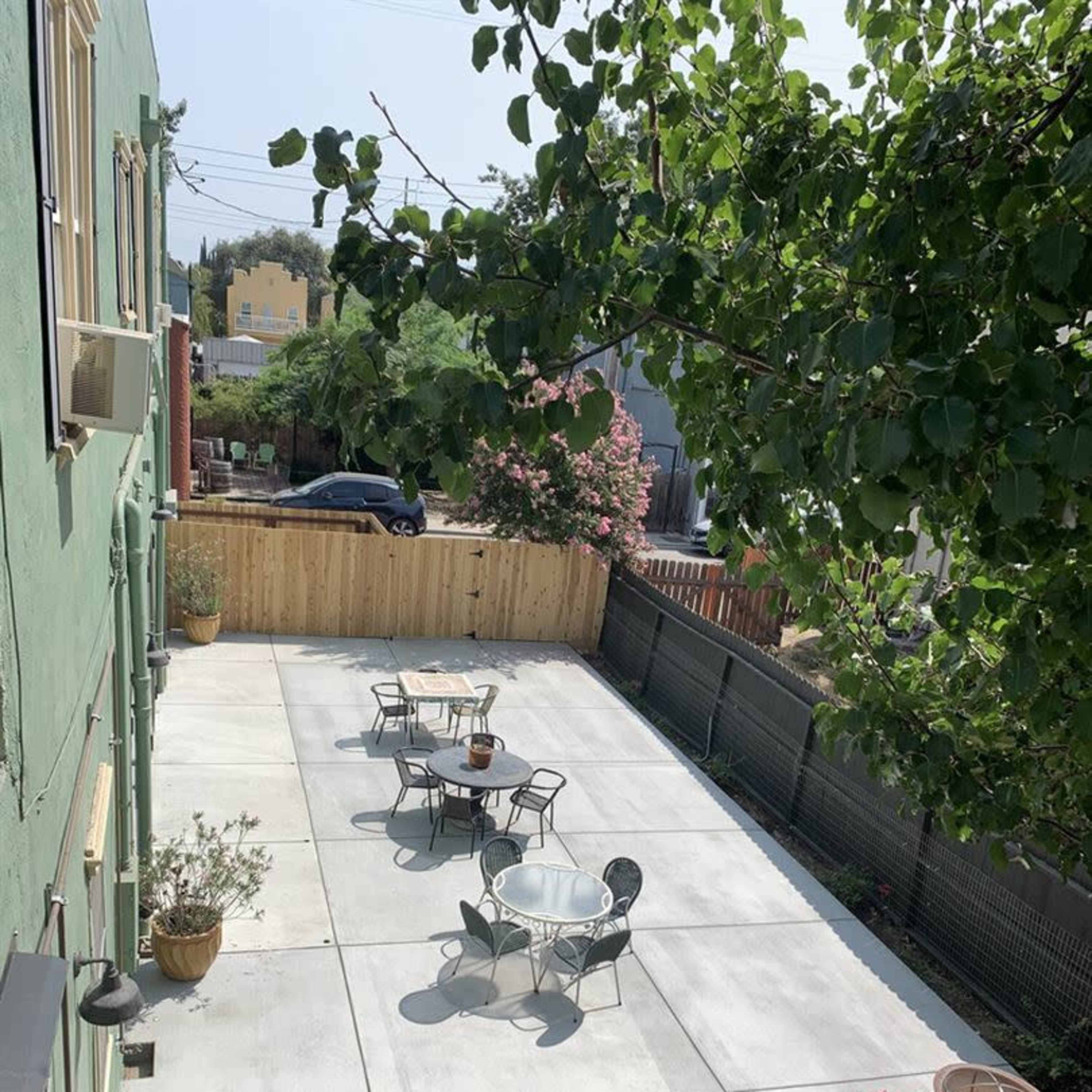 A concrete patio features several round tables and chairs surrounded by greenery and a wooden fence.