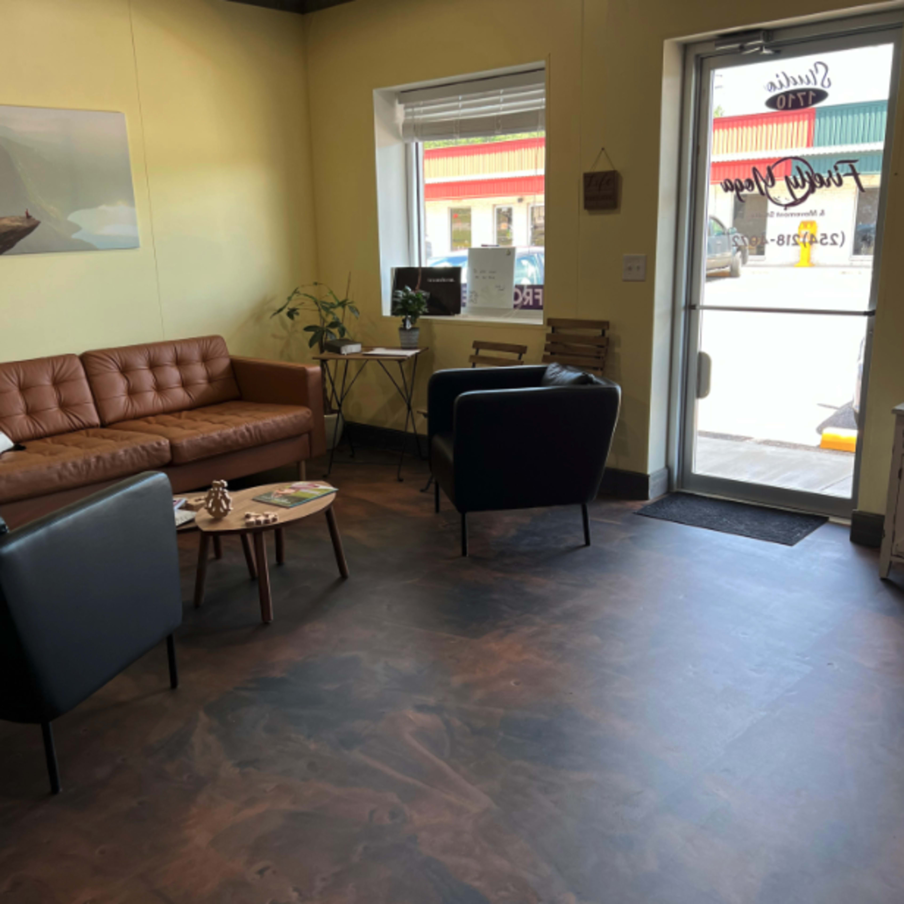 A cozy waiting area with a brown leather couch, two black chairs, a small table, and a door leading outside.