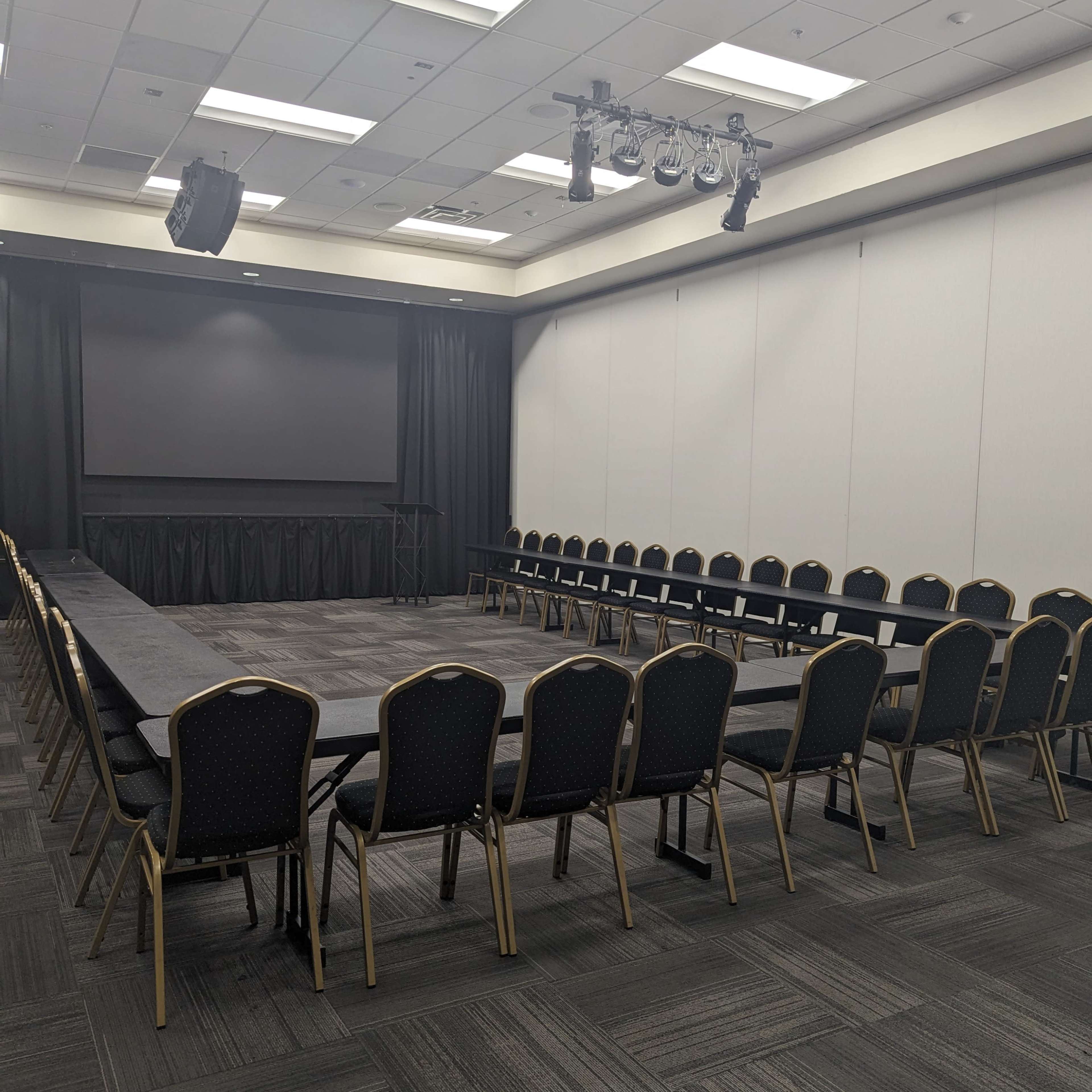 A conference room arranged with a large round table surrounded by chairs, facing a screen at the front.