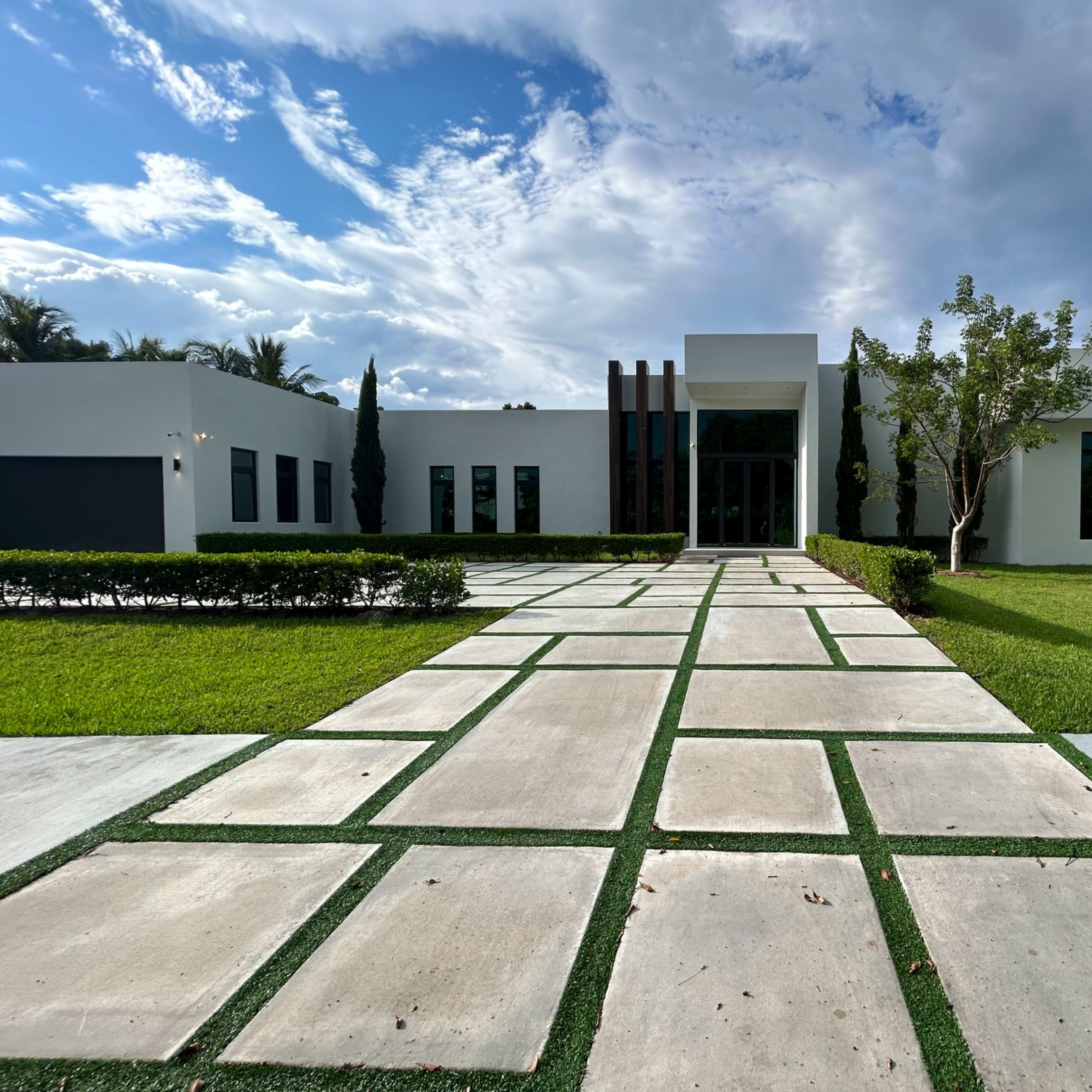 A modern house with a paved pathway and neatly trimmed landscaping leads to the entrance, set against a backdrop of cloudy skies.
