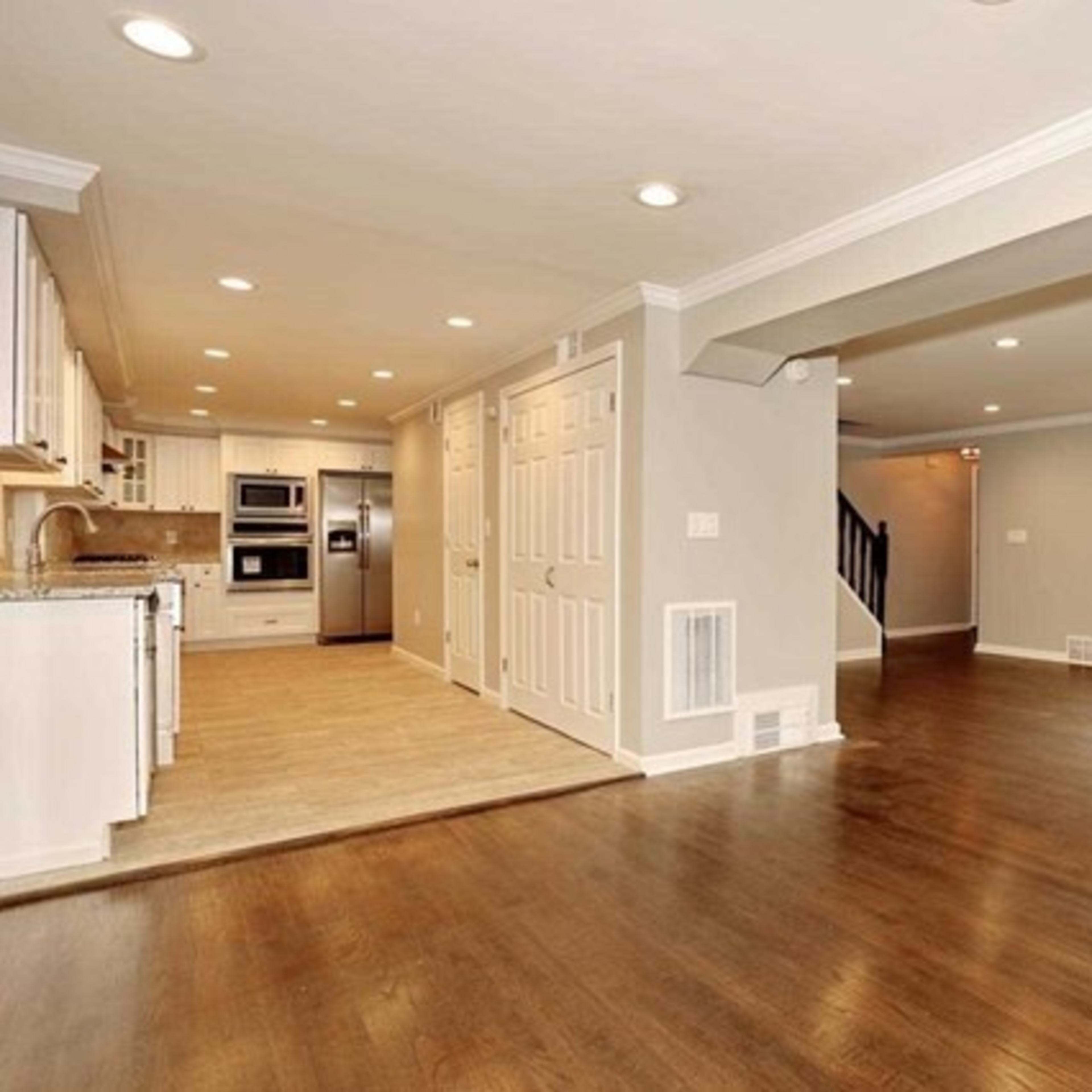 A spacious, open-concept living area with hardwood floors, a large window, and a modern kitchen featuring white cabinetry and stainless steel appliances.