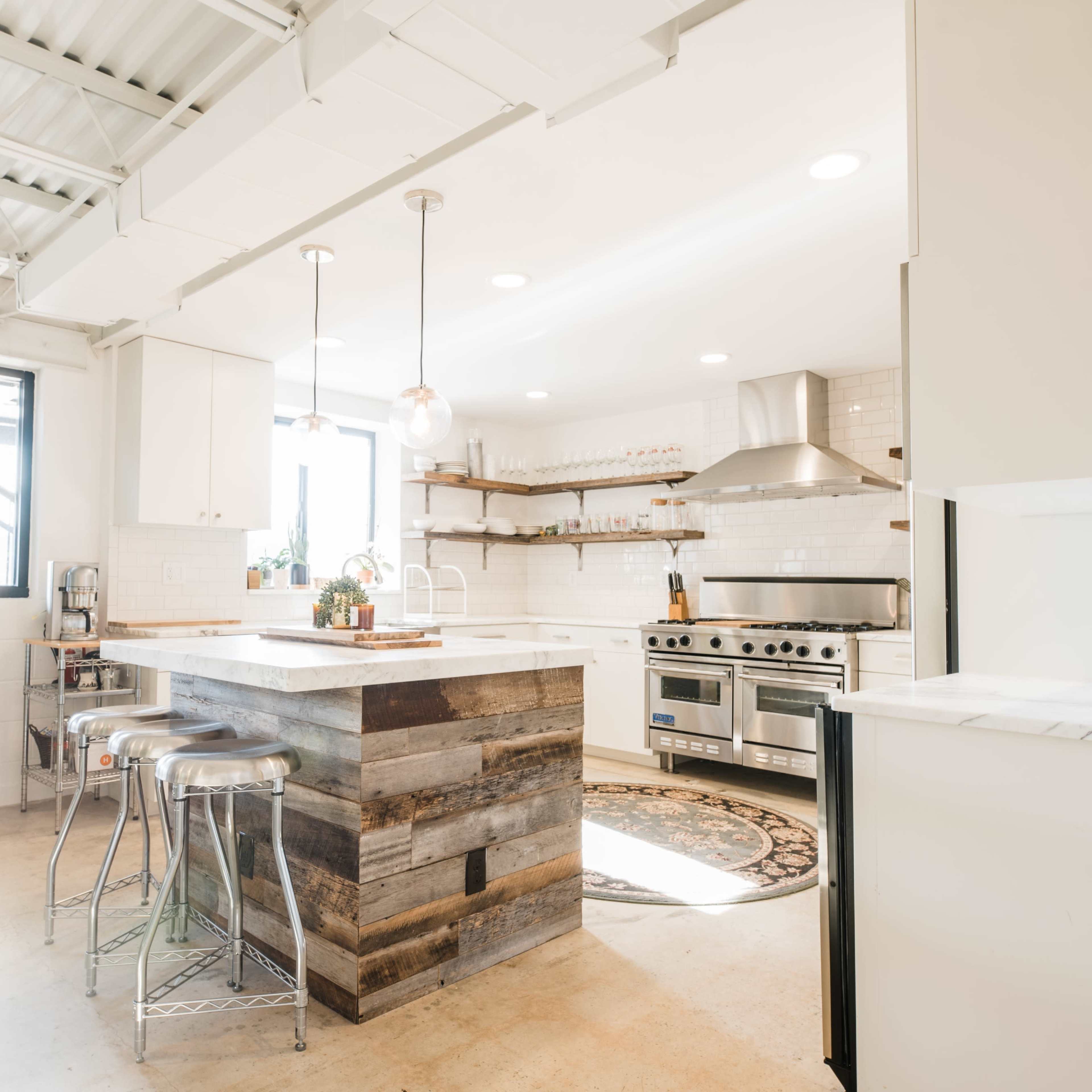A modern kitchen features a spacious island made of reclaimed wood, stainless steel appliances, and large windows that allow natural light to enter.