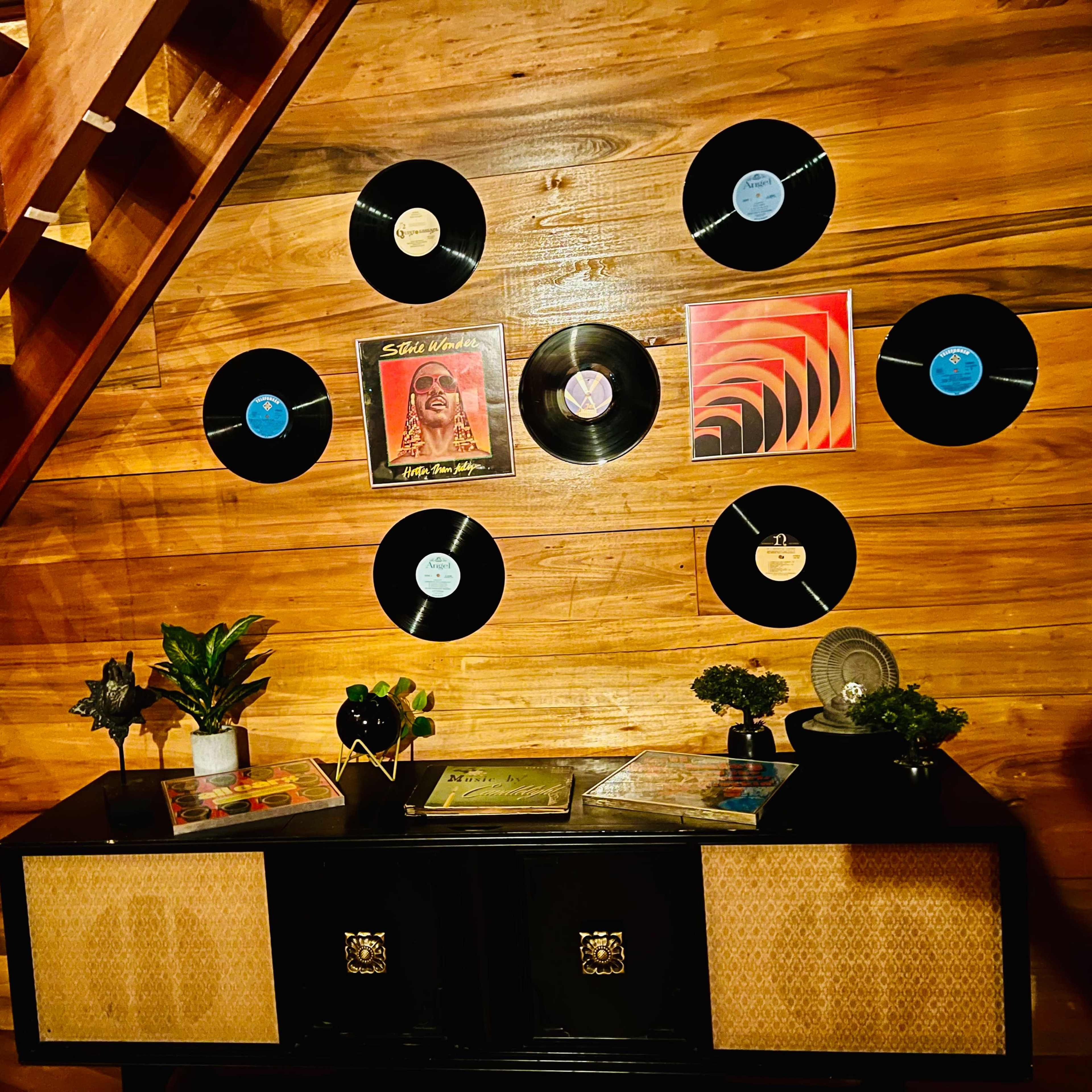 A wooden staircase leads to a wall decorated with vinyl records above a dark cabinet topped with plants and magazines.