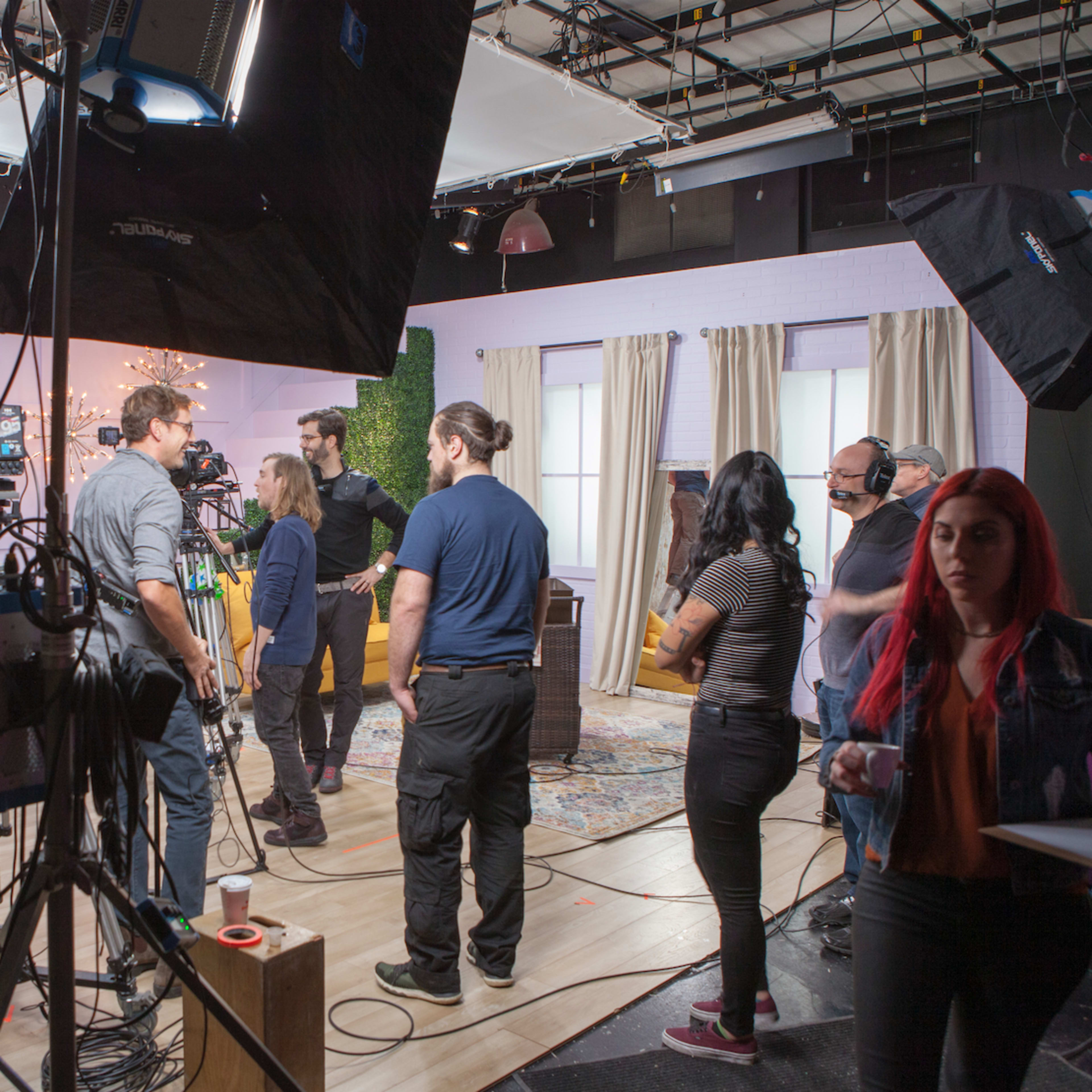A film crew prepares for a shoot in a television studio with cameras, lighting equipment, and a backdrop arrangement.
