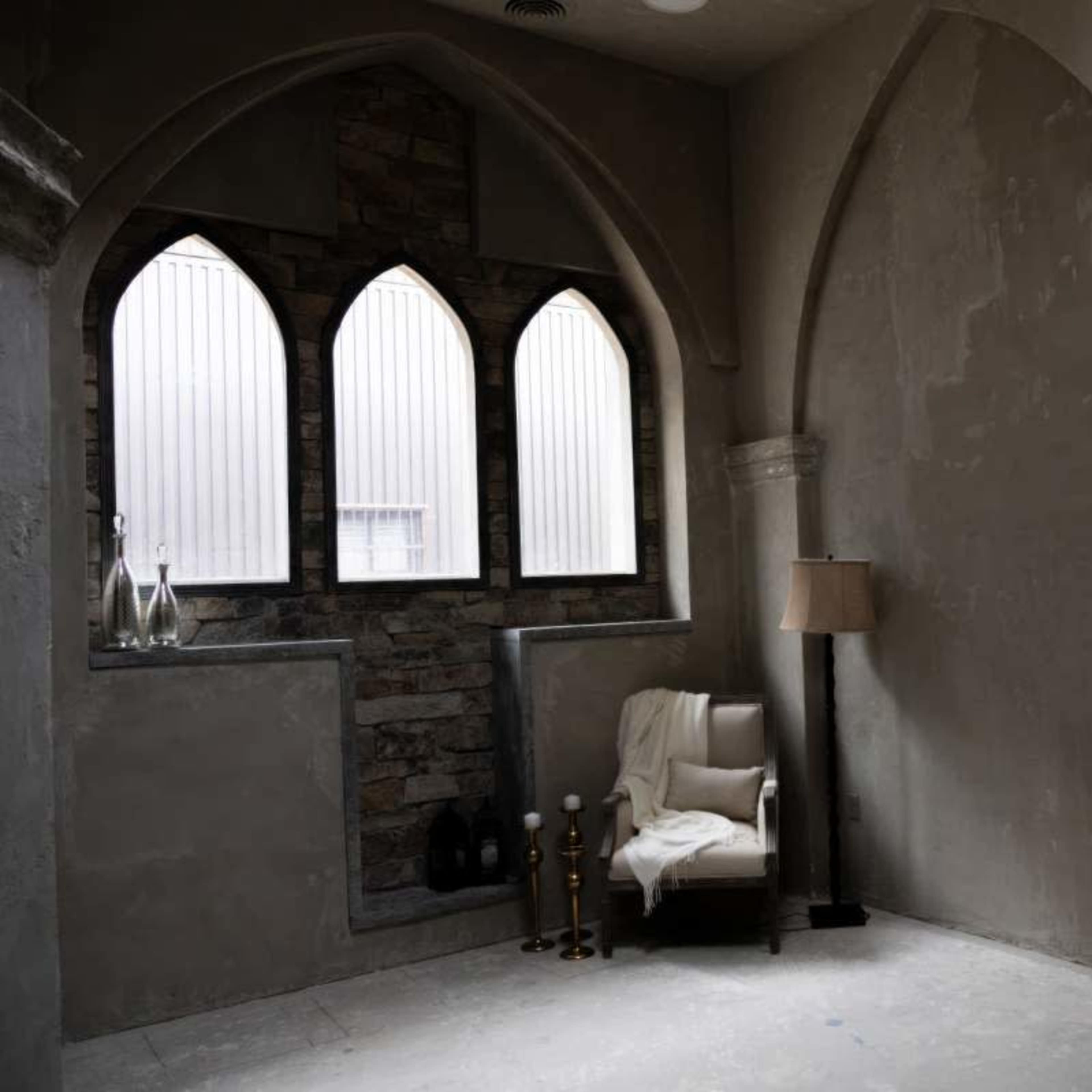 An interior corner of a room featuring a chair beside a lamp, with arched windows and stone accents on the walls.
