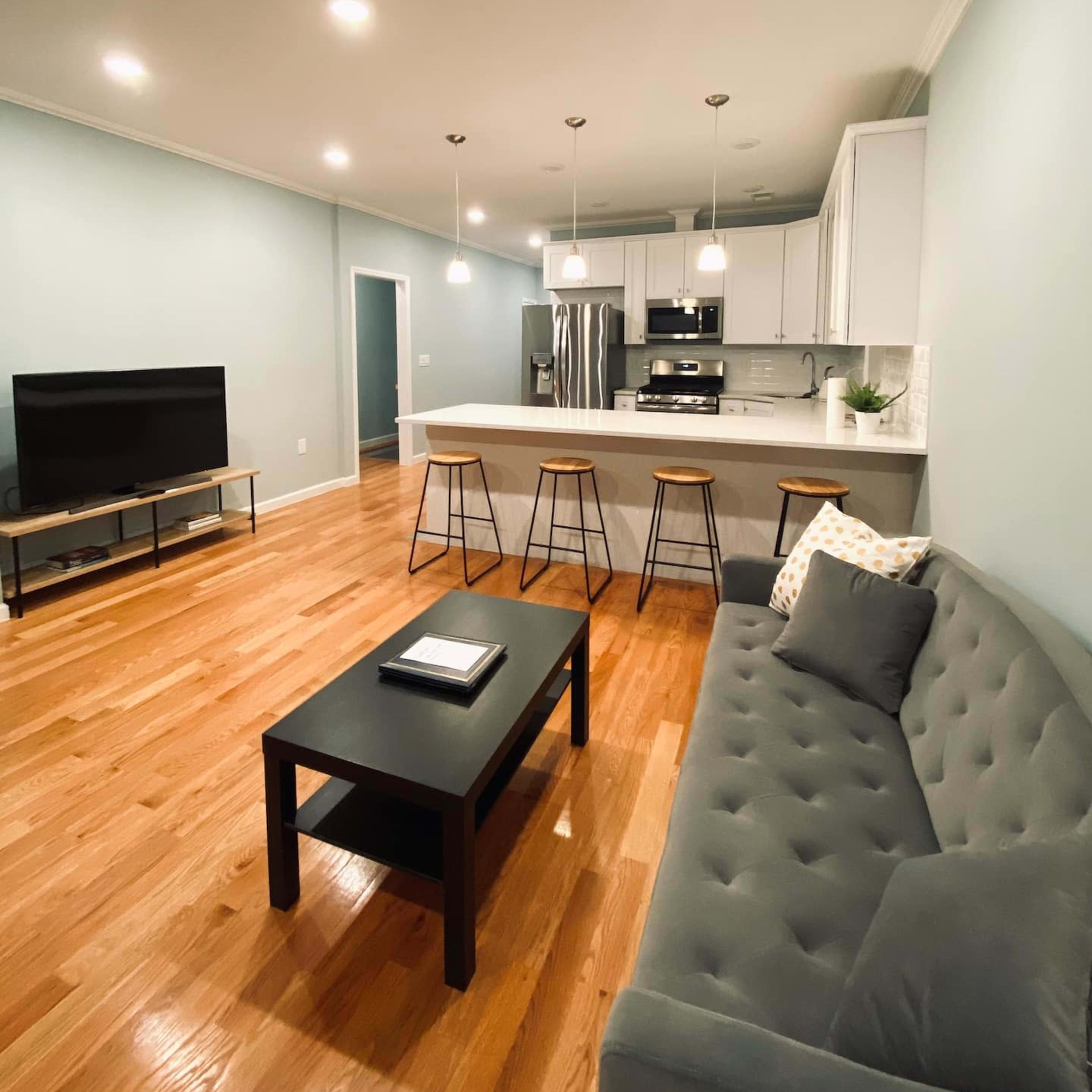 A modern living room and kitchen area featuring a gray couch, a black coffee table, bar stools at a kitchen island, and a large television on a stand.