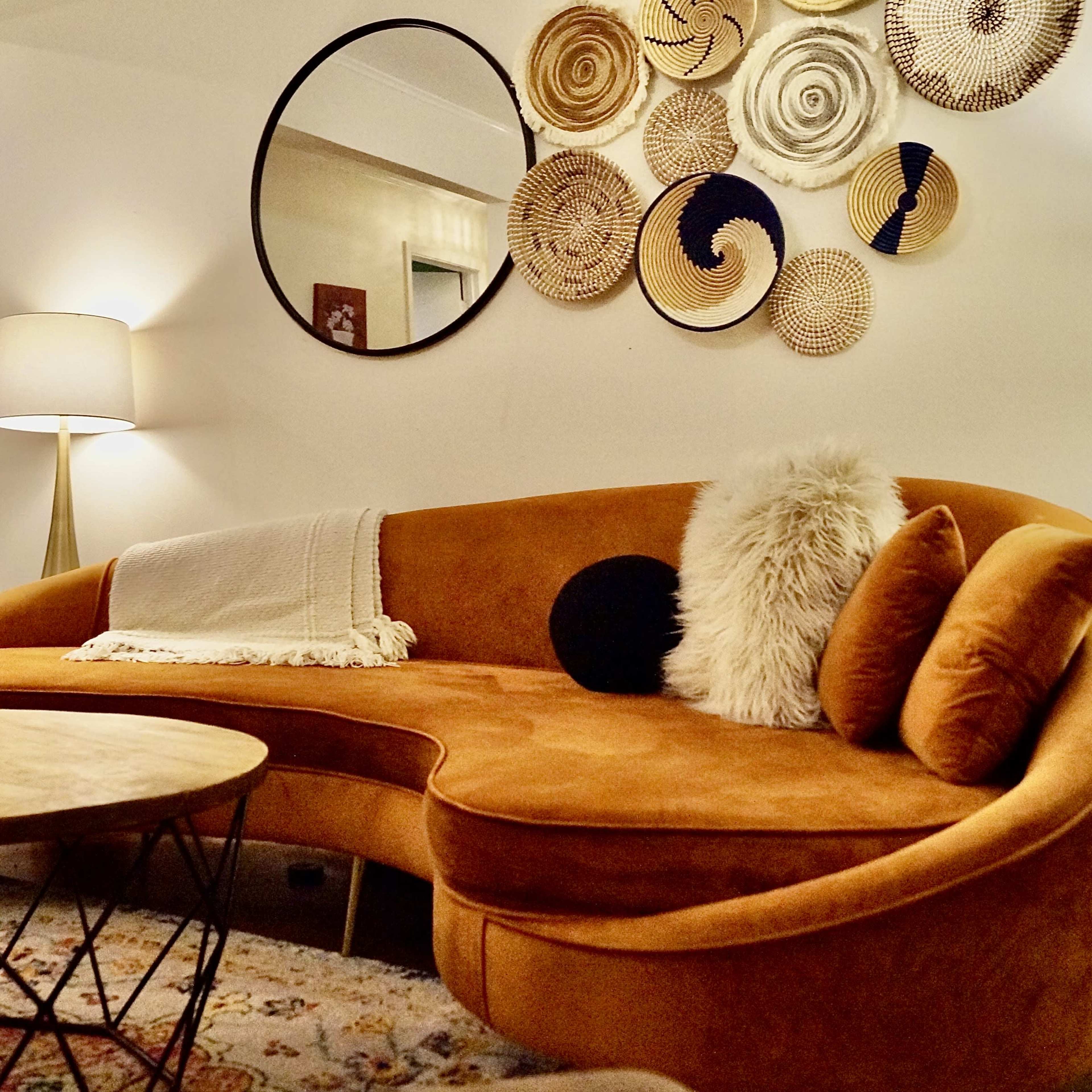 A modern living room featuring an orange curved sofa, a round mirror, decorative wall hangings, and a wooden coffee table.