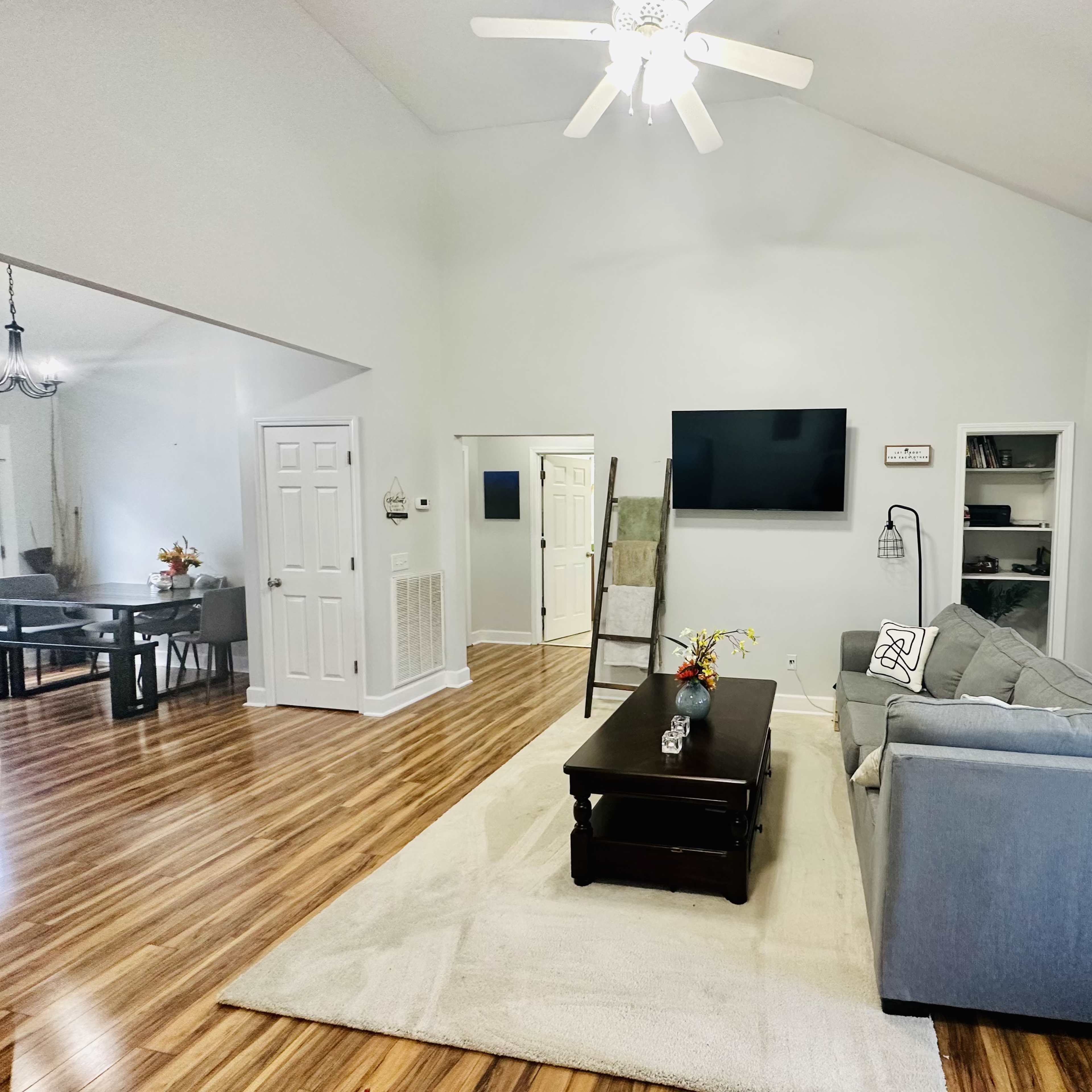 A spacious living room with a gray sofa, a dark coffee table, and a flat-screen television mounted on a light-colored wall.