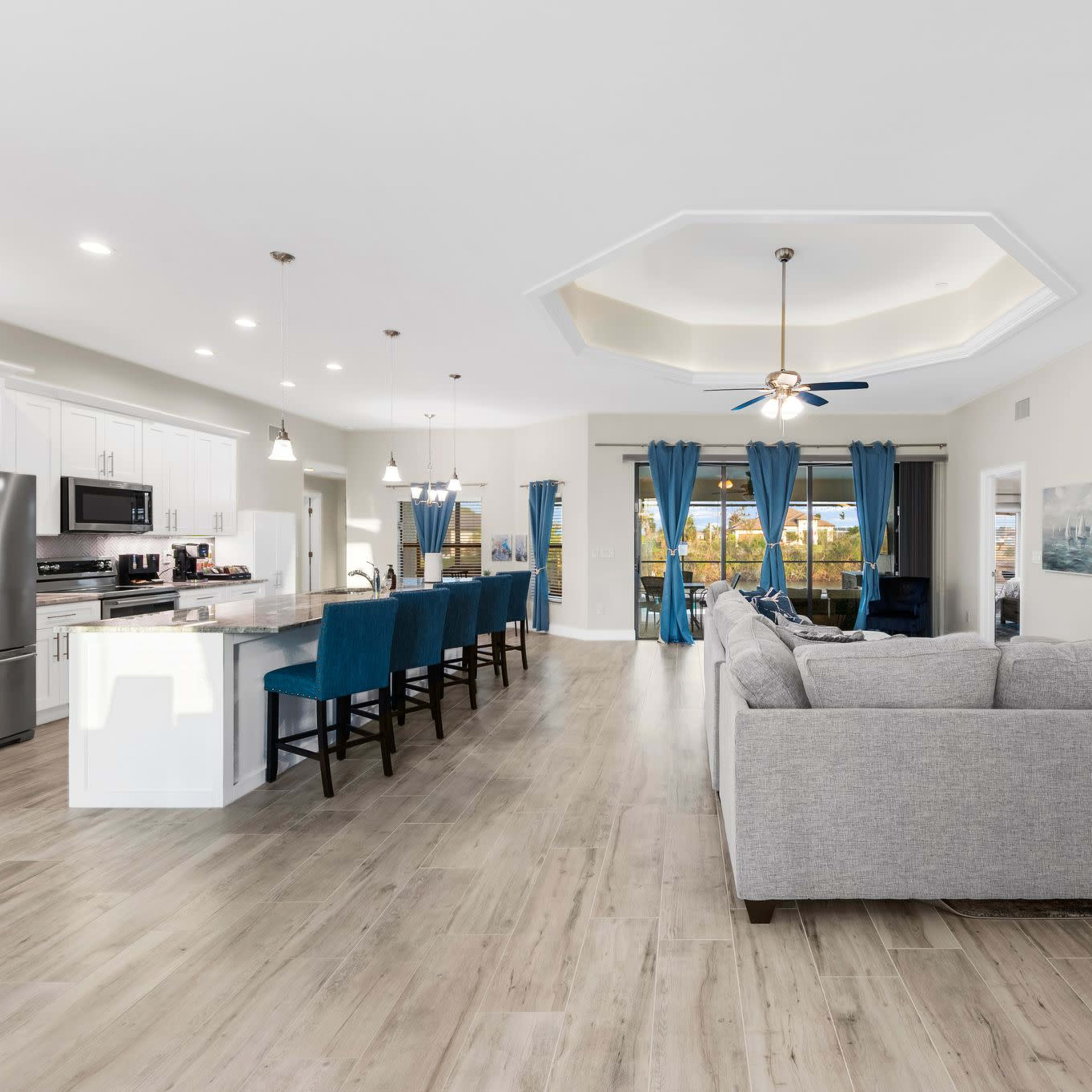 A bright, open living space with a gray sectional sofa, a kitchen with white cabinets, and blue bar stools.