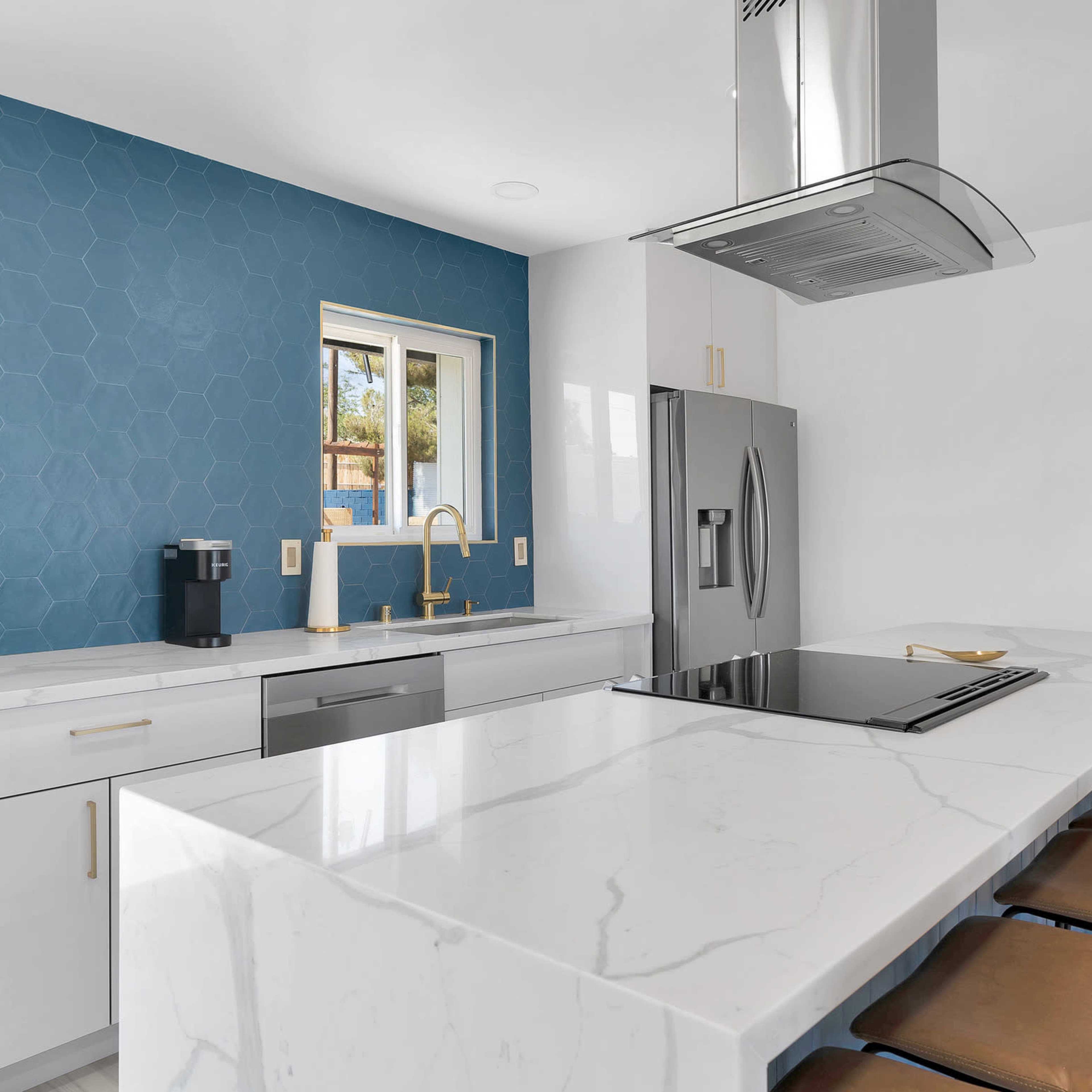 A modern kitchen features a blue hexagonal tiled wall, white marble countertops, and stainless steel appliances.