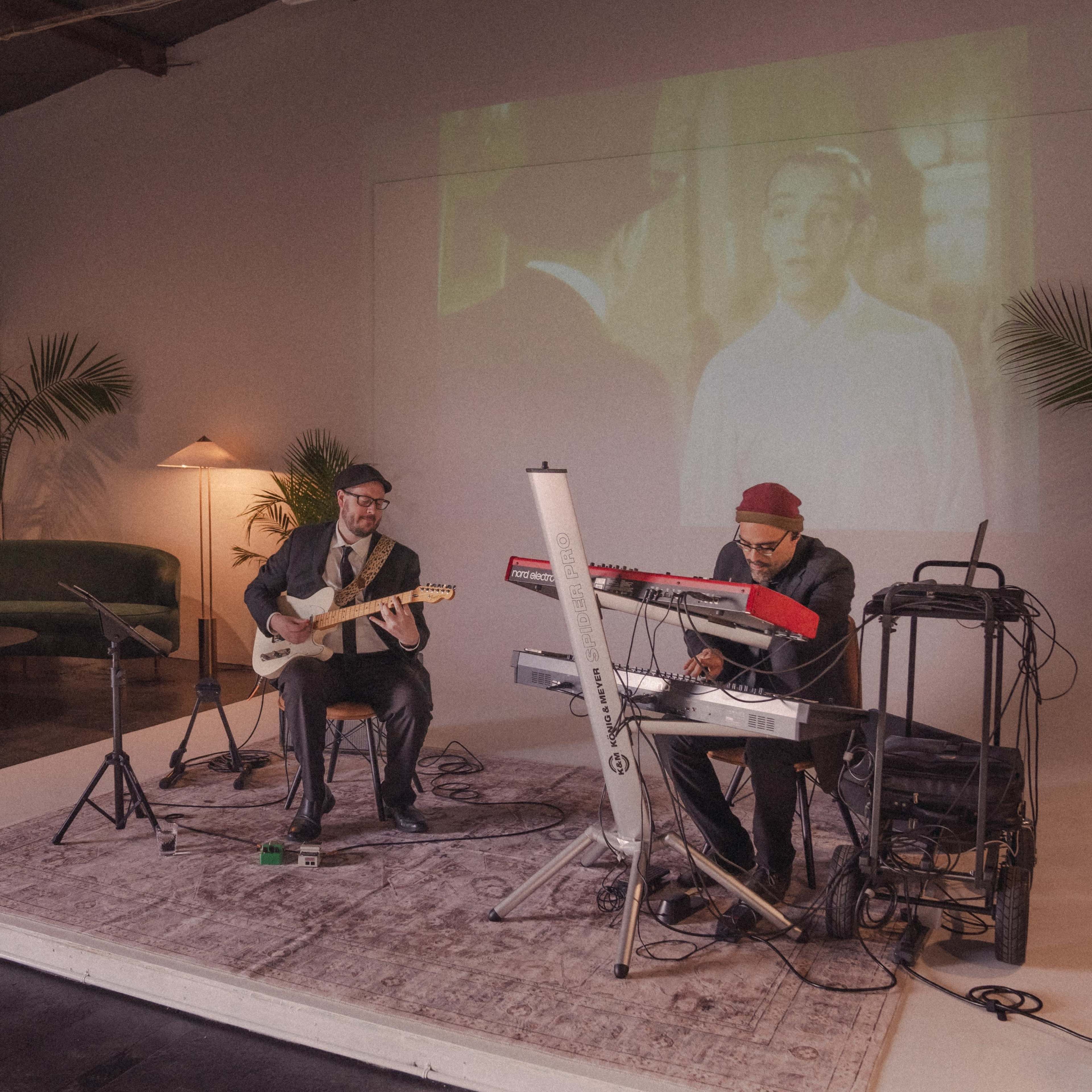 Two musicians perform on a small stage in a modern space with a projected film in the background.