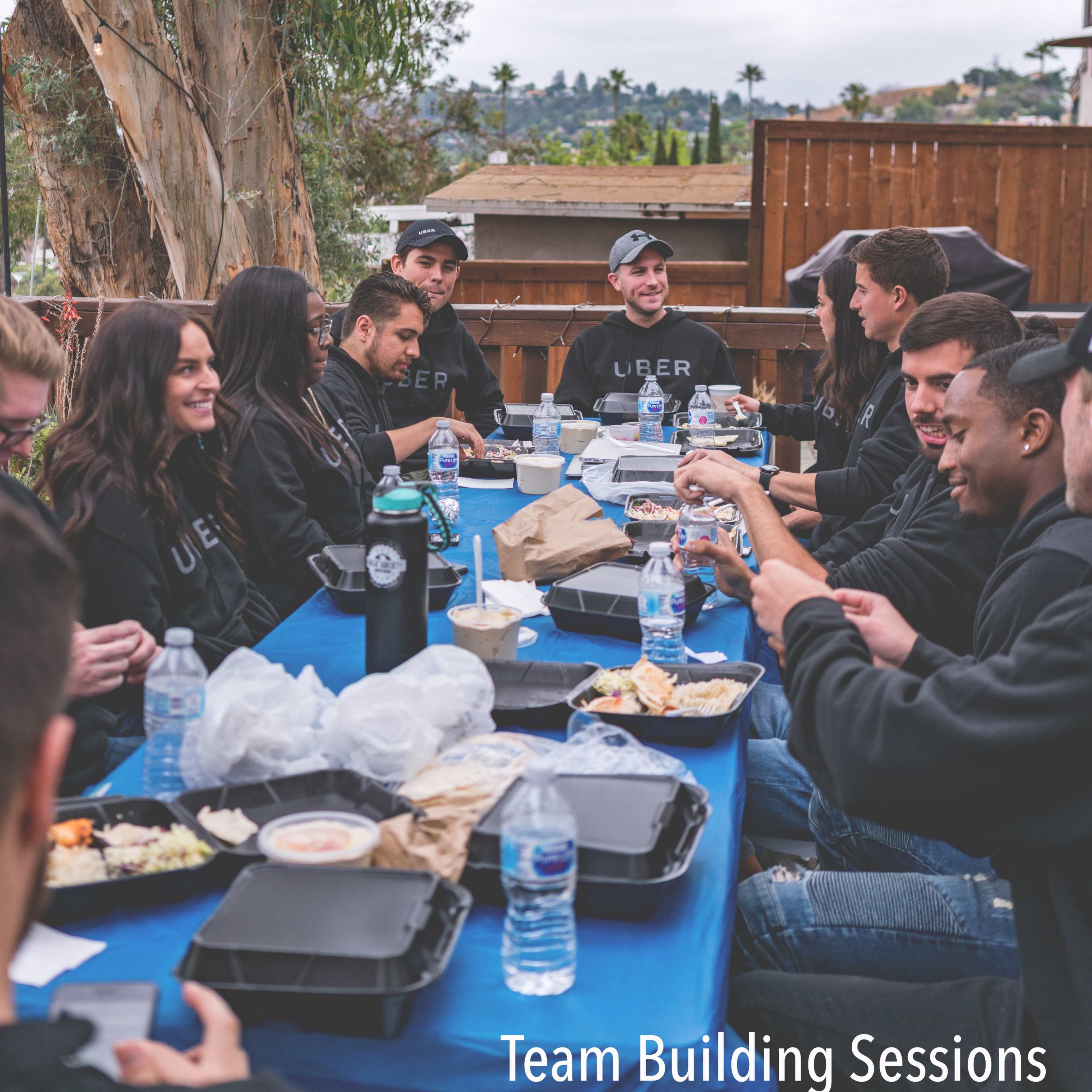 A group of people is seated around a table outdoors, sharing food and beverages during a team-building session.
