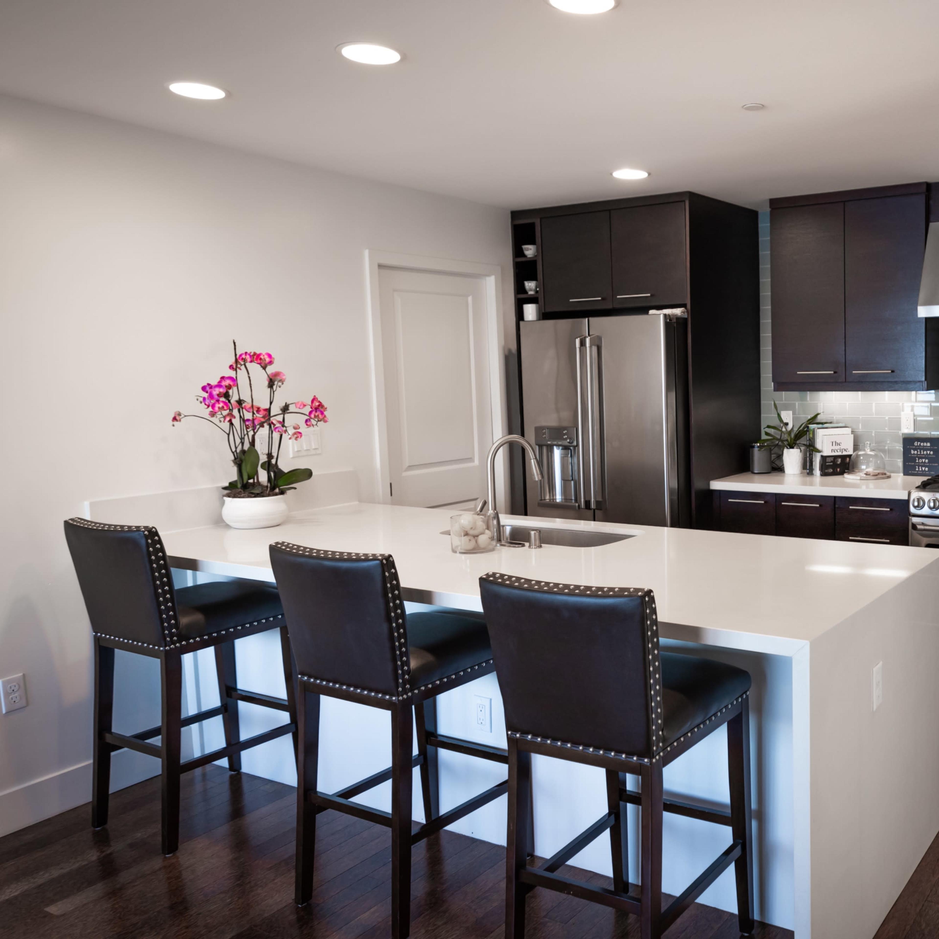 A modern kitchen with dark cabinetry, stainless steel appliances, a white countertop, and three black barstools at a kitchen island.