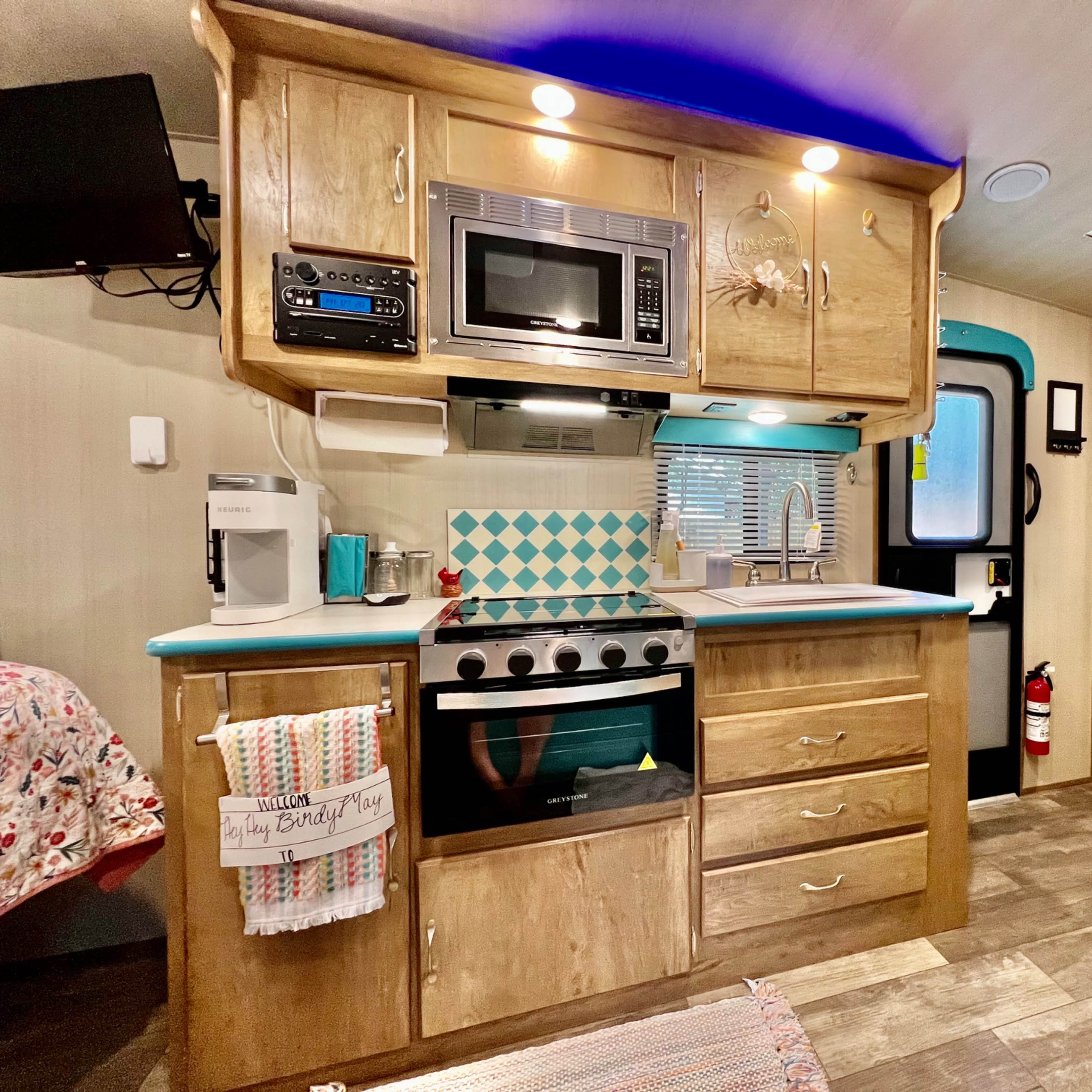 A compact kitchen area in an RV, featuring cabinetry, a stove, a microwave, and a sink, with blue and white tiled accents.