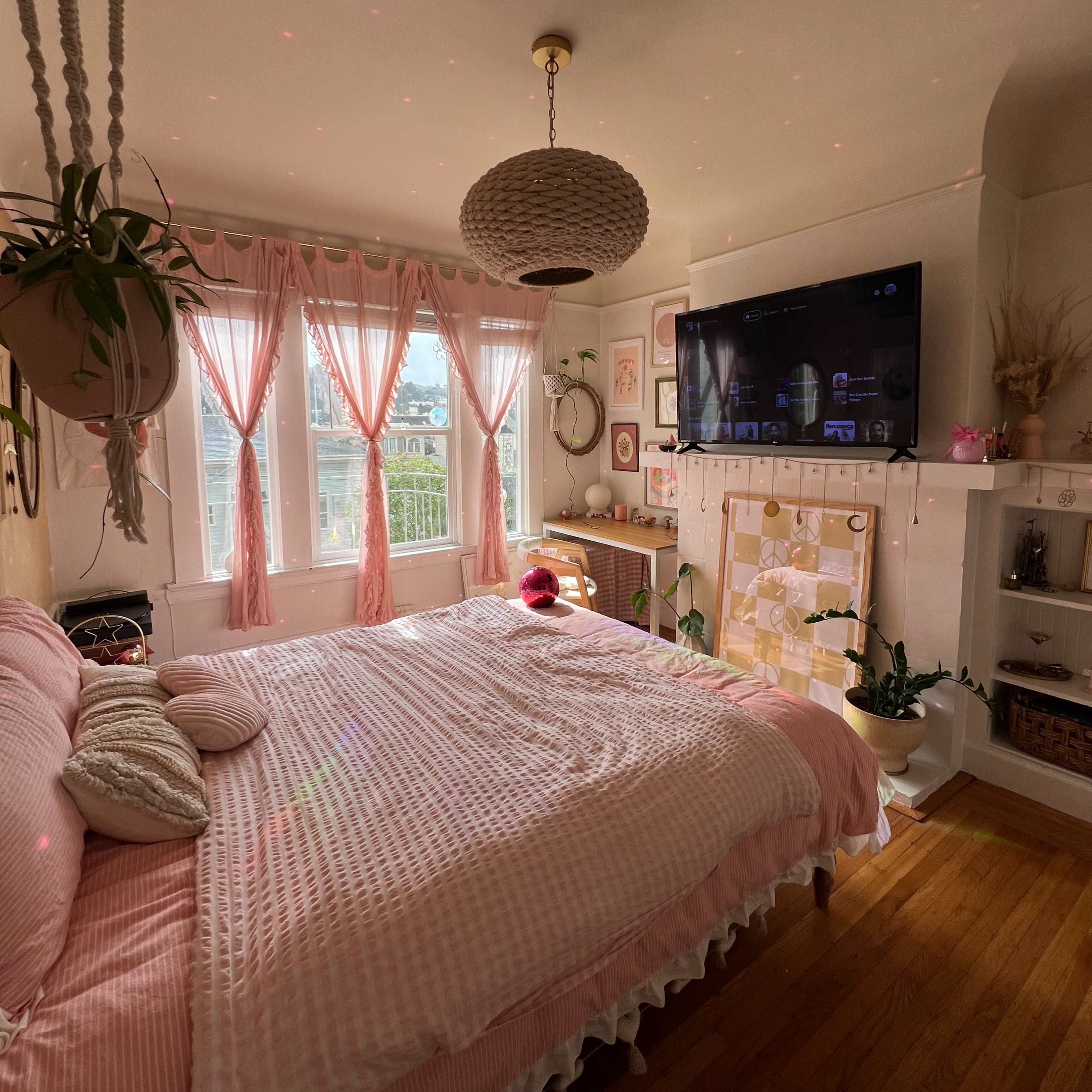A bright bedroom with a large bed covered in pink bedding, windows draped with light pink curtains, and a television mounted on the wall above a decorative shelf.