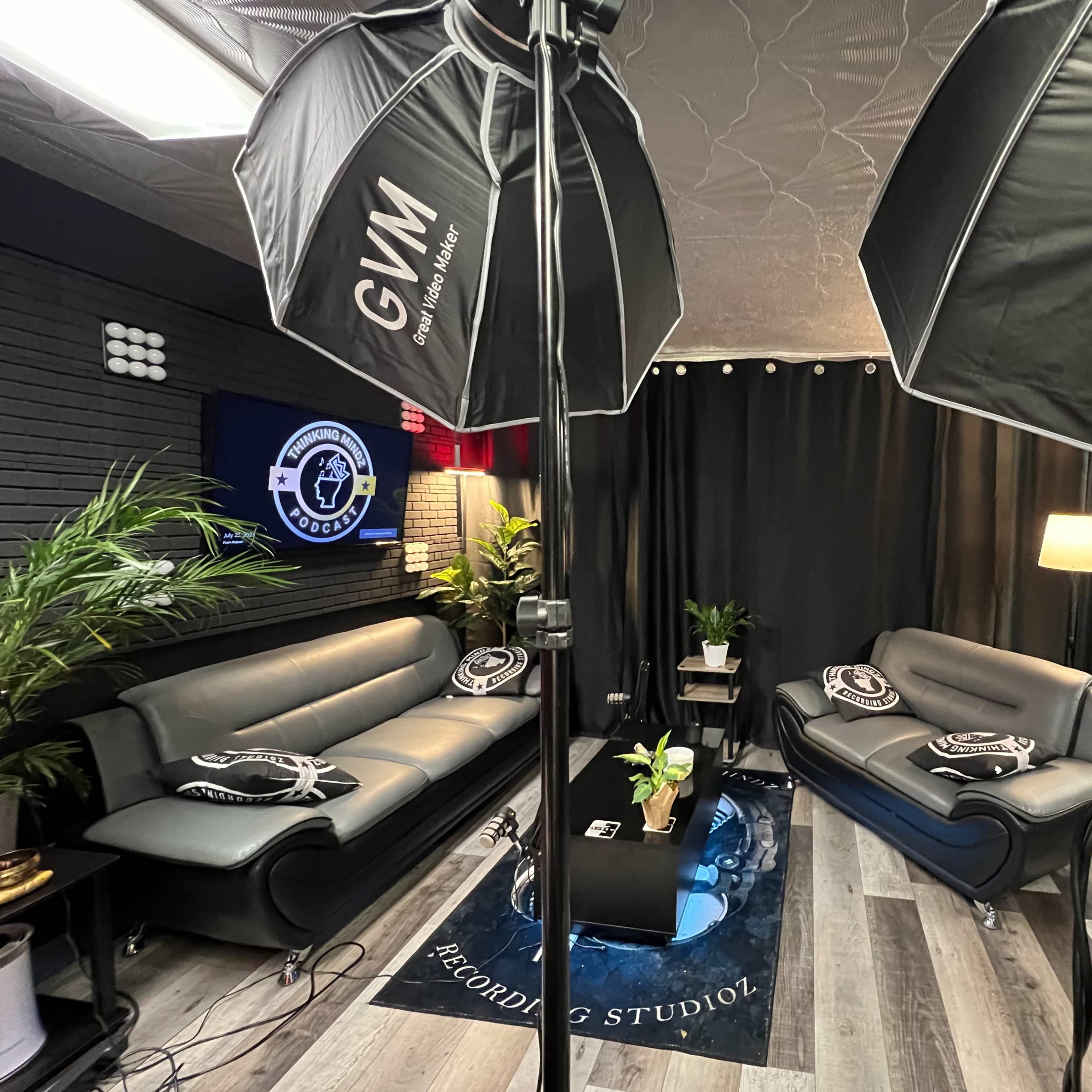 A recording studio with two black sofas, a coffee table, and lighting equipment set up for a shoot, surrounded by black curtains and decorative plants.