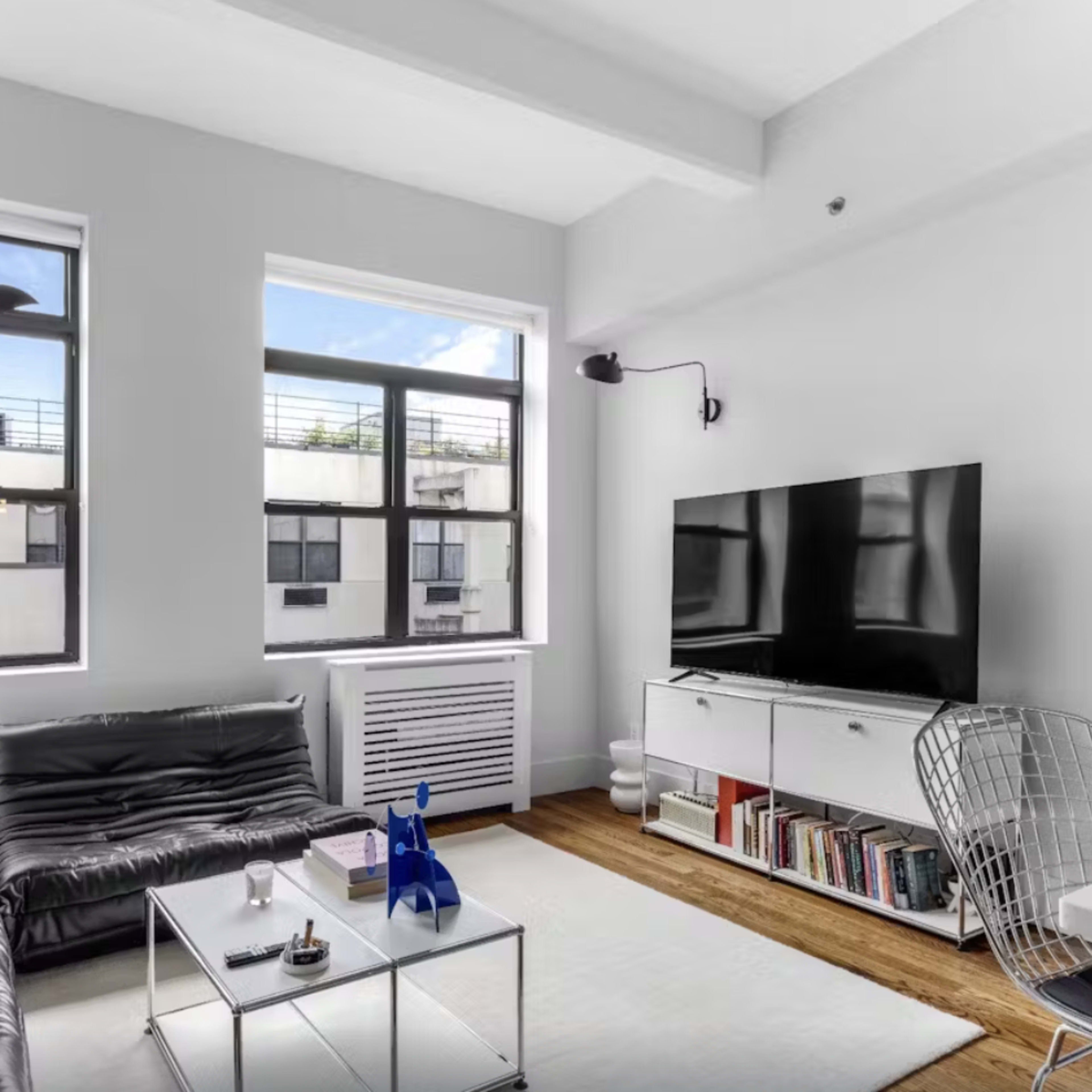 A modern living room with a black leather sofa, a white coffee table, a television mounted on the wall, and large windows letting in natural light.
