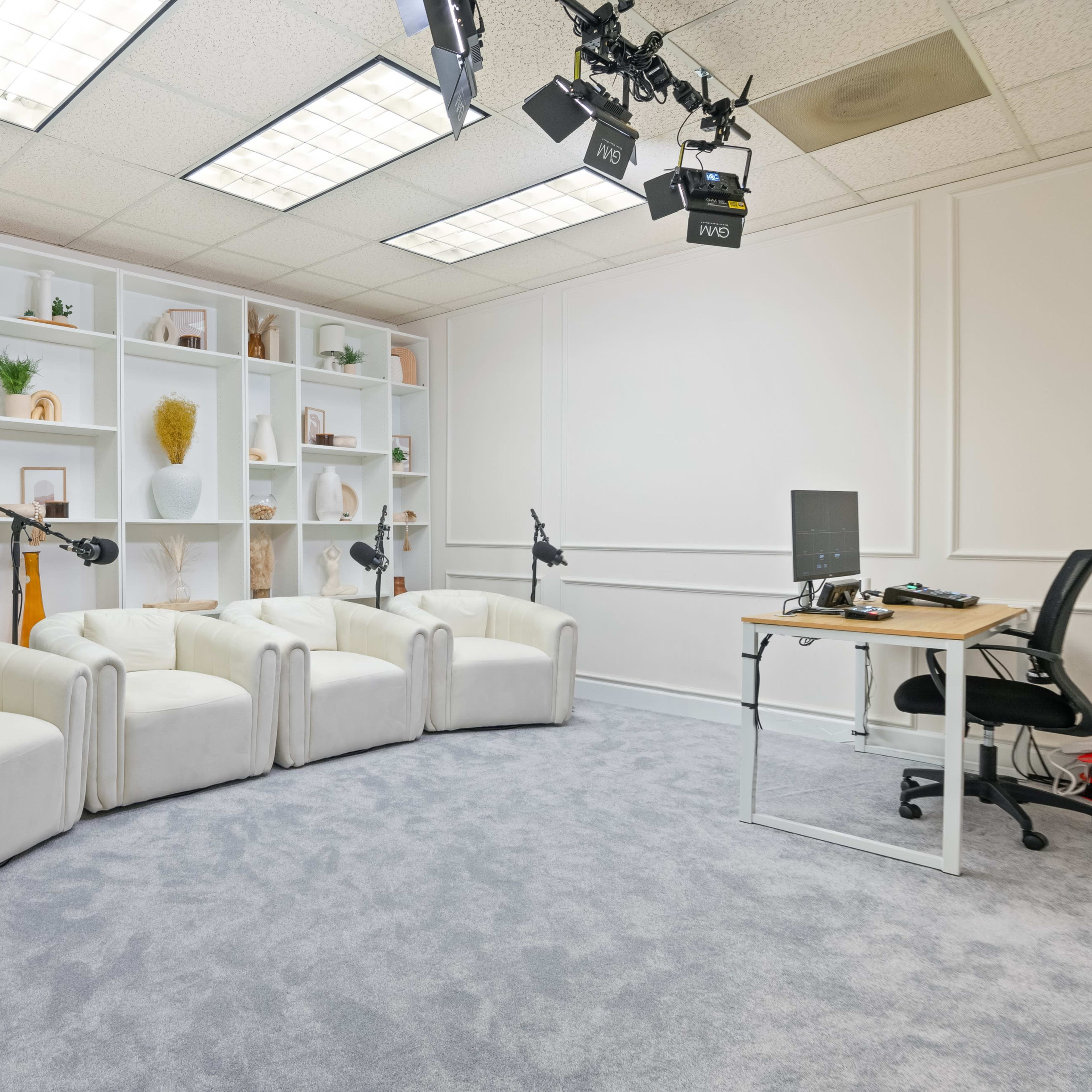 A modern interview set features three white chairs arranged in front of a bookshelf, with microphones on stands, a desk, and a chair in a well-lit room.