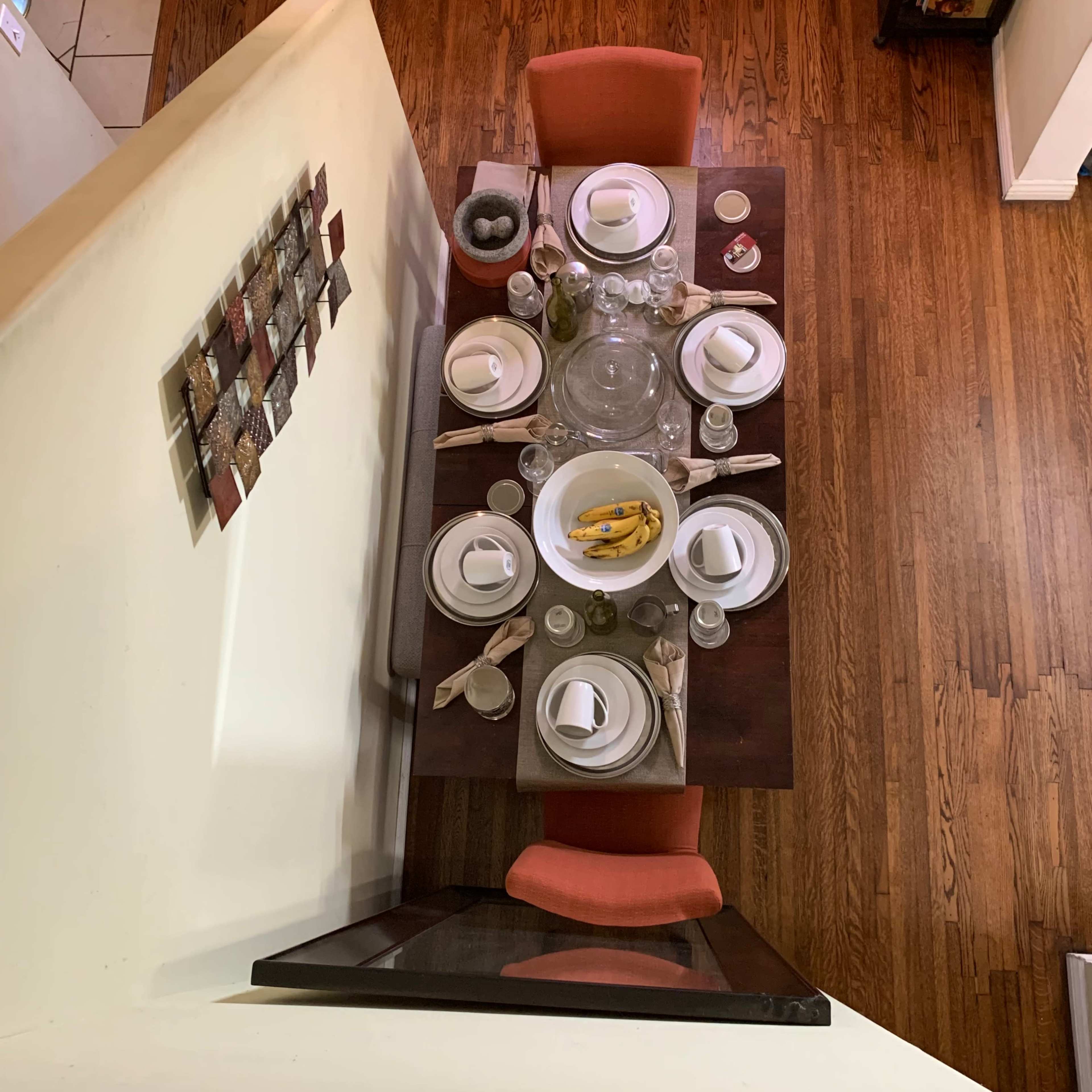 A dining table set for a meal, featuring plates, glasses, utensils, and a centerpiece bowl with bananas, surrounded by orange chairs.
