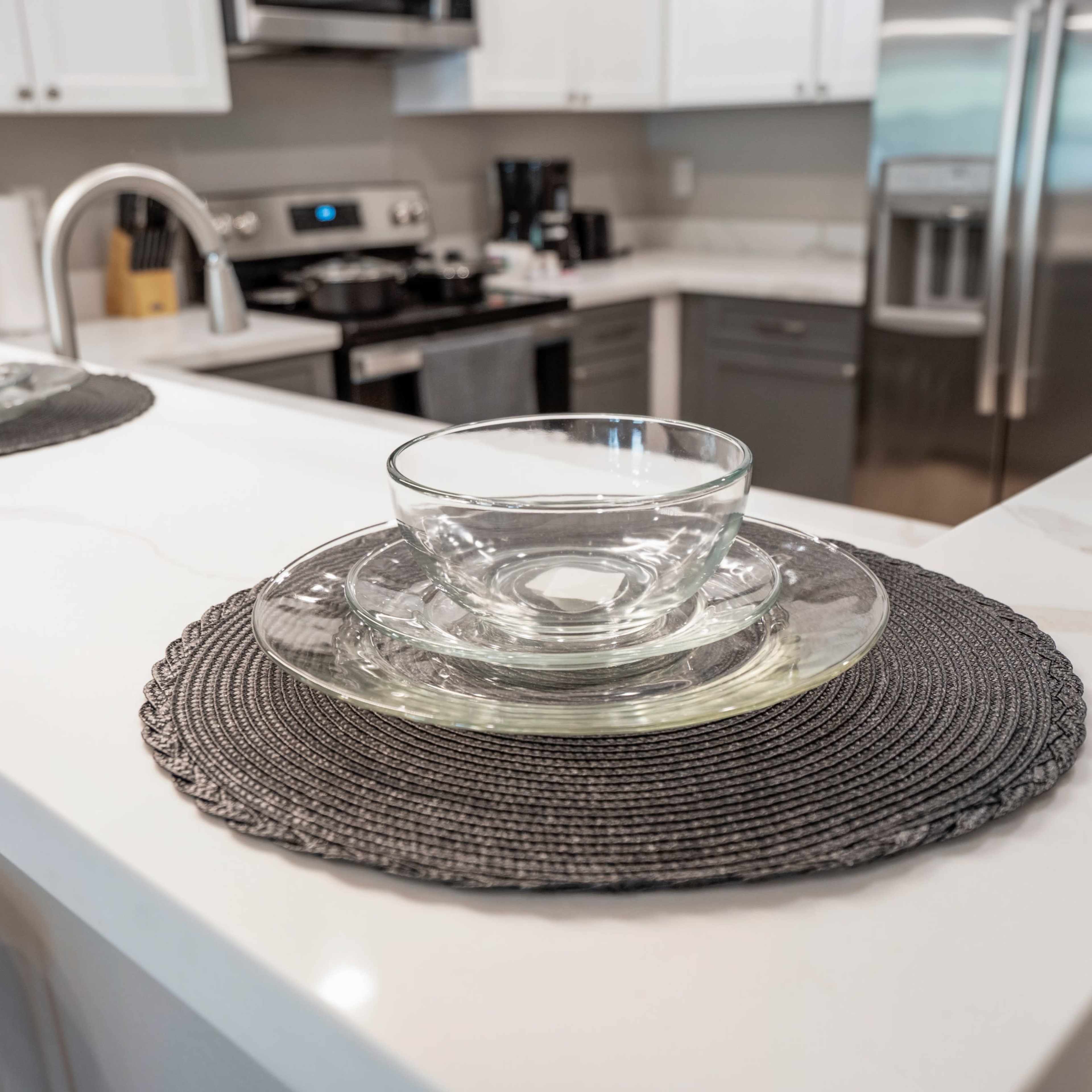 A kitchen countertop features a glass bowl and plate set on a woven placemat, with stainless steel appliances visible in the background.