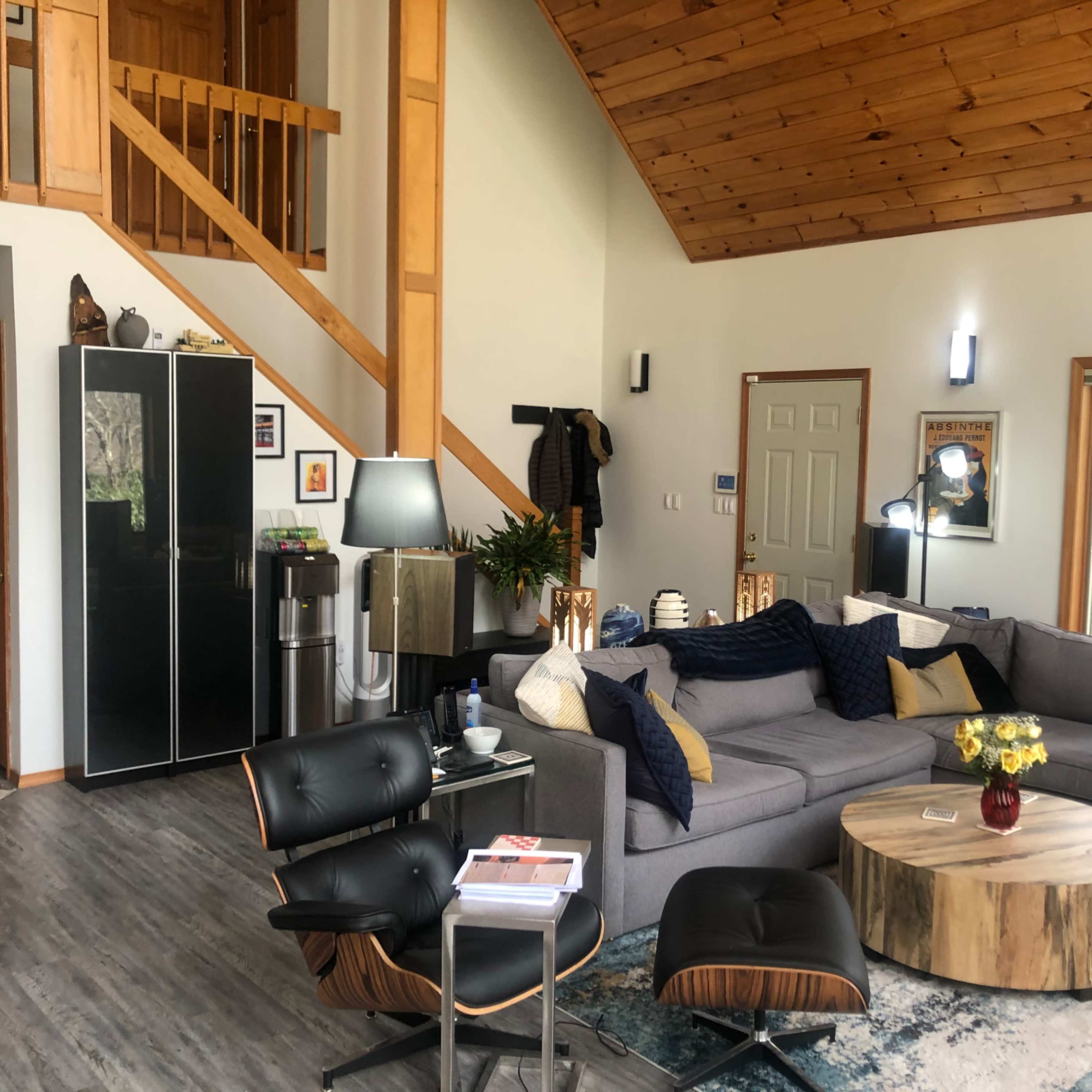 The scene shows a modern living room with a gray sectional sofa, a wooden coffee table, and a staircase leading to an upper level, complemented by large windows that allow natural light.