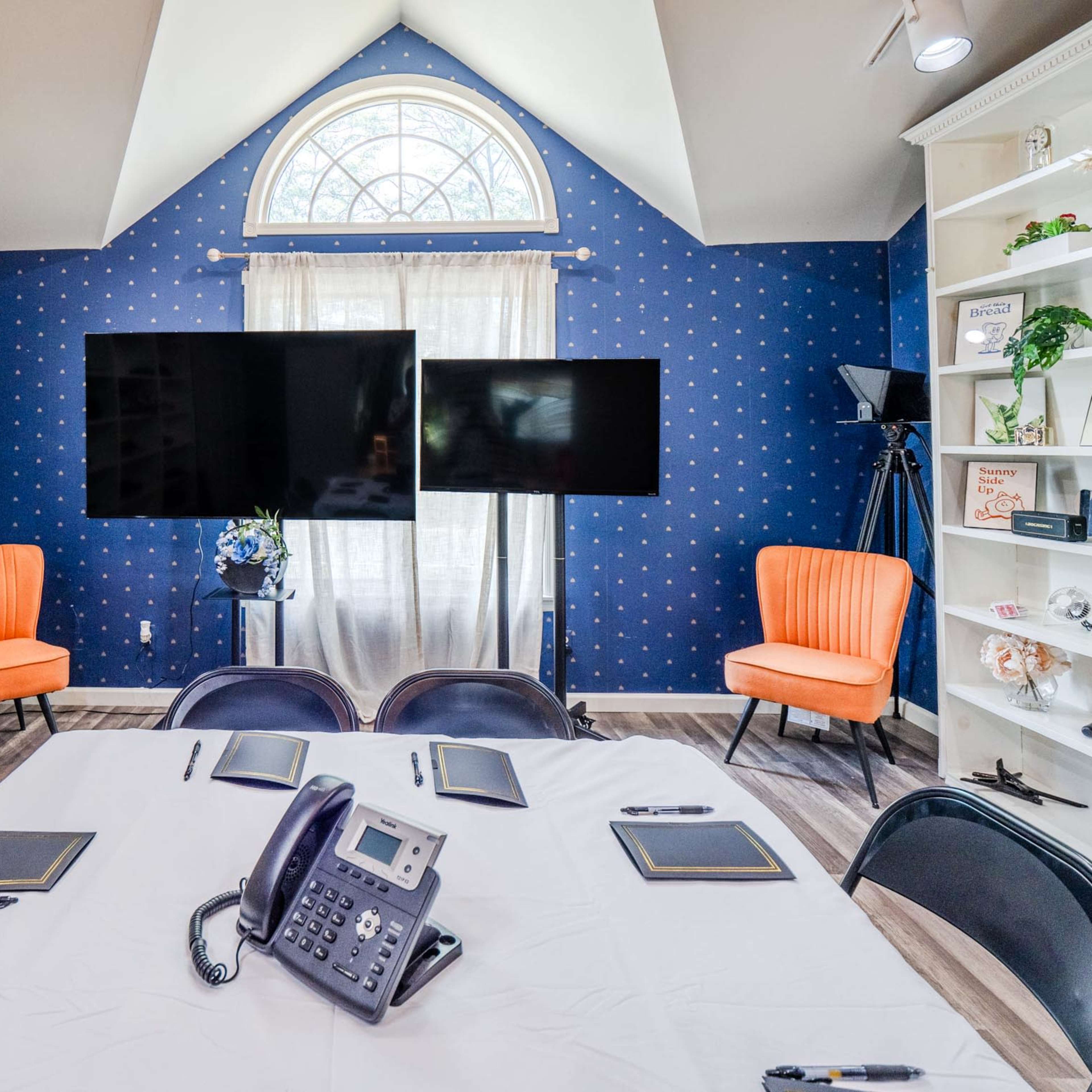 A conference room features a round table with notepads and a phone, flanked by two orange chairs, two television screens, and a large window.