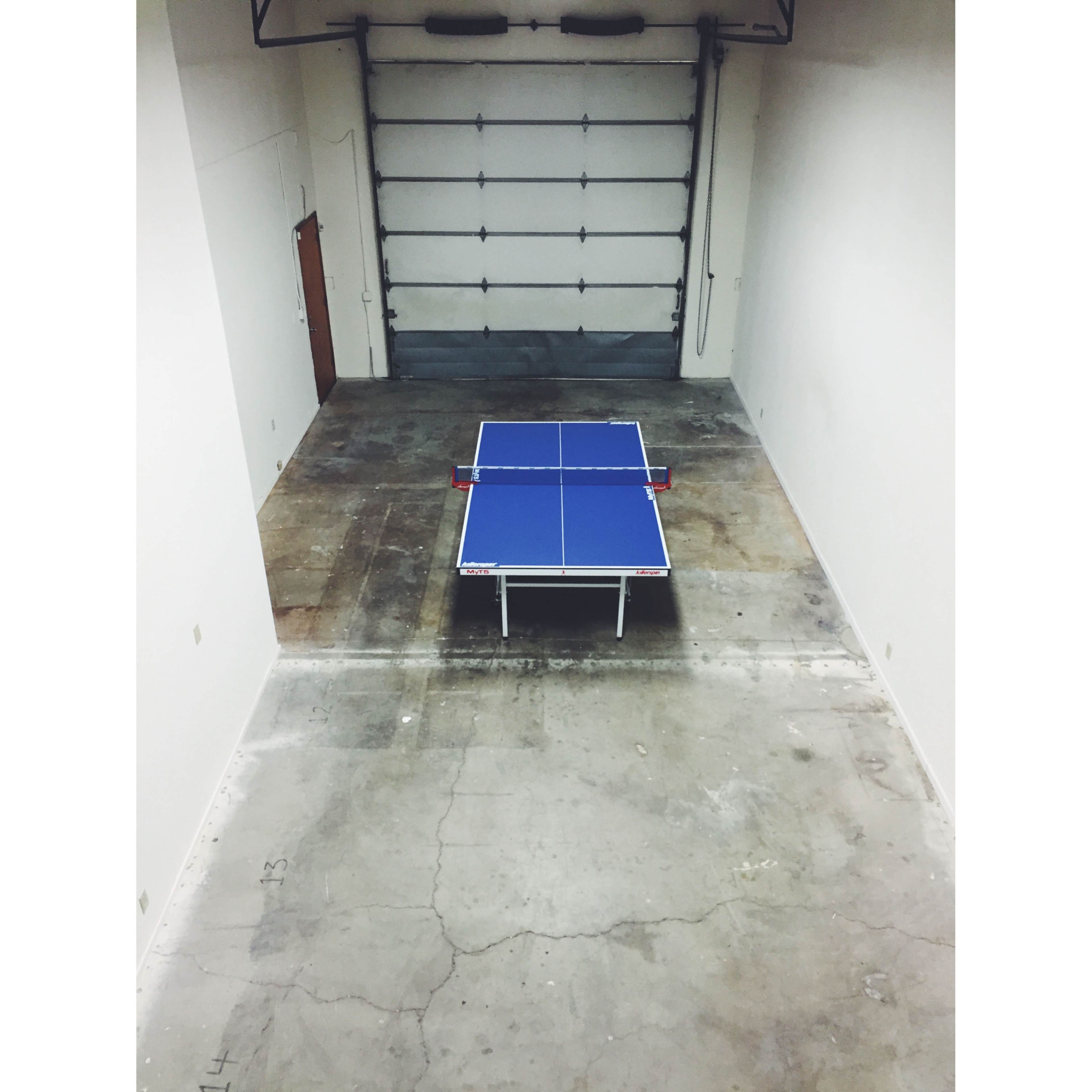 A blue ping pong table is positioned in the center of a large, empty room with concrete floors and white walls.