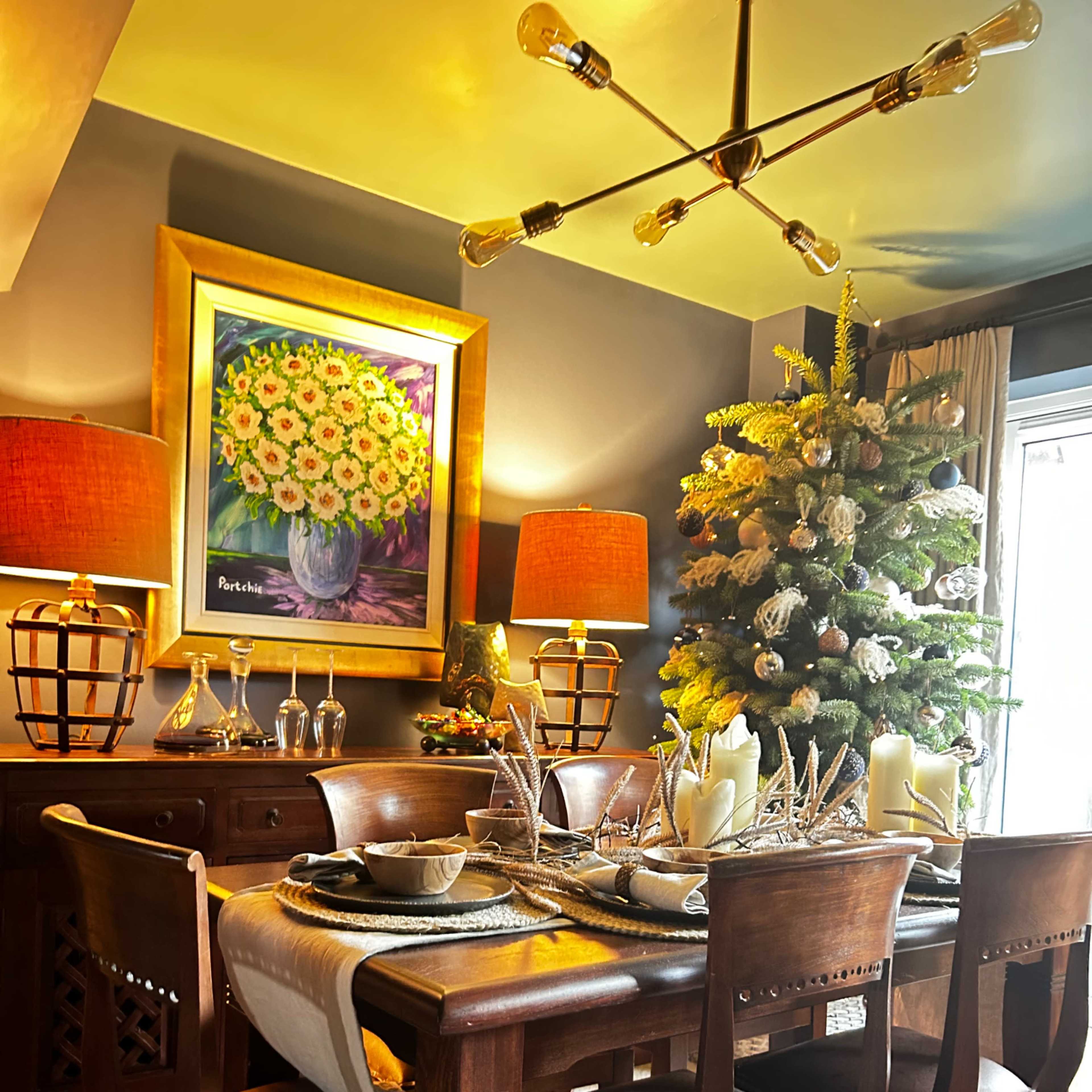 A dining room features a wooden table set with dishes and candles, surrounded by chairs, against a backdrop of a decorated Christmas tree and a large floral painting.