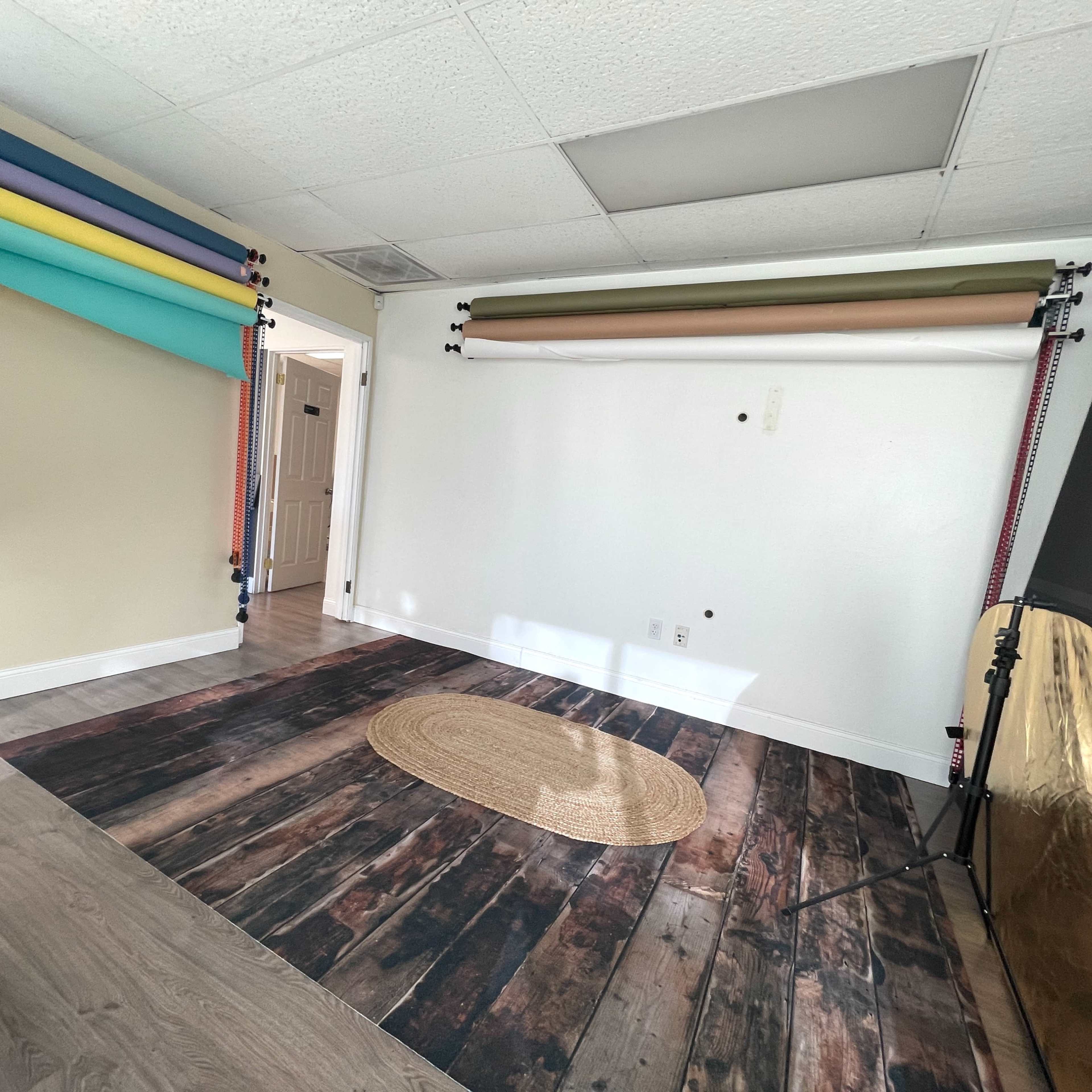 A photo studio with a white wall, a round rug on a wooden floor, and several color backdrop rolls hanging from the ceiling.
