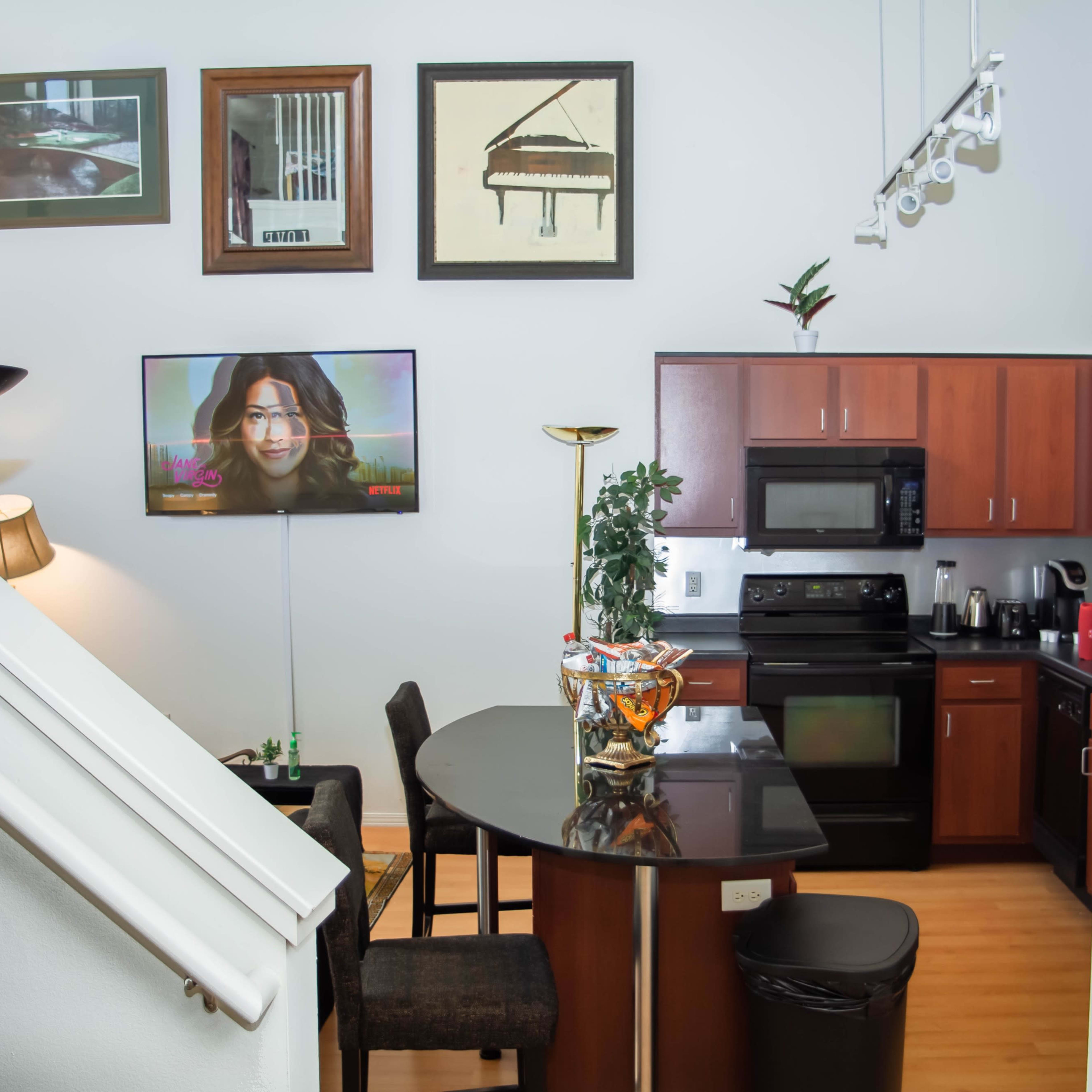 A modern kitchen with dark wood cabinetry and appliances, a round table with chairs, and a living area featuring a television displaying a woman.