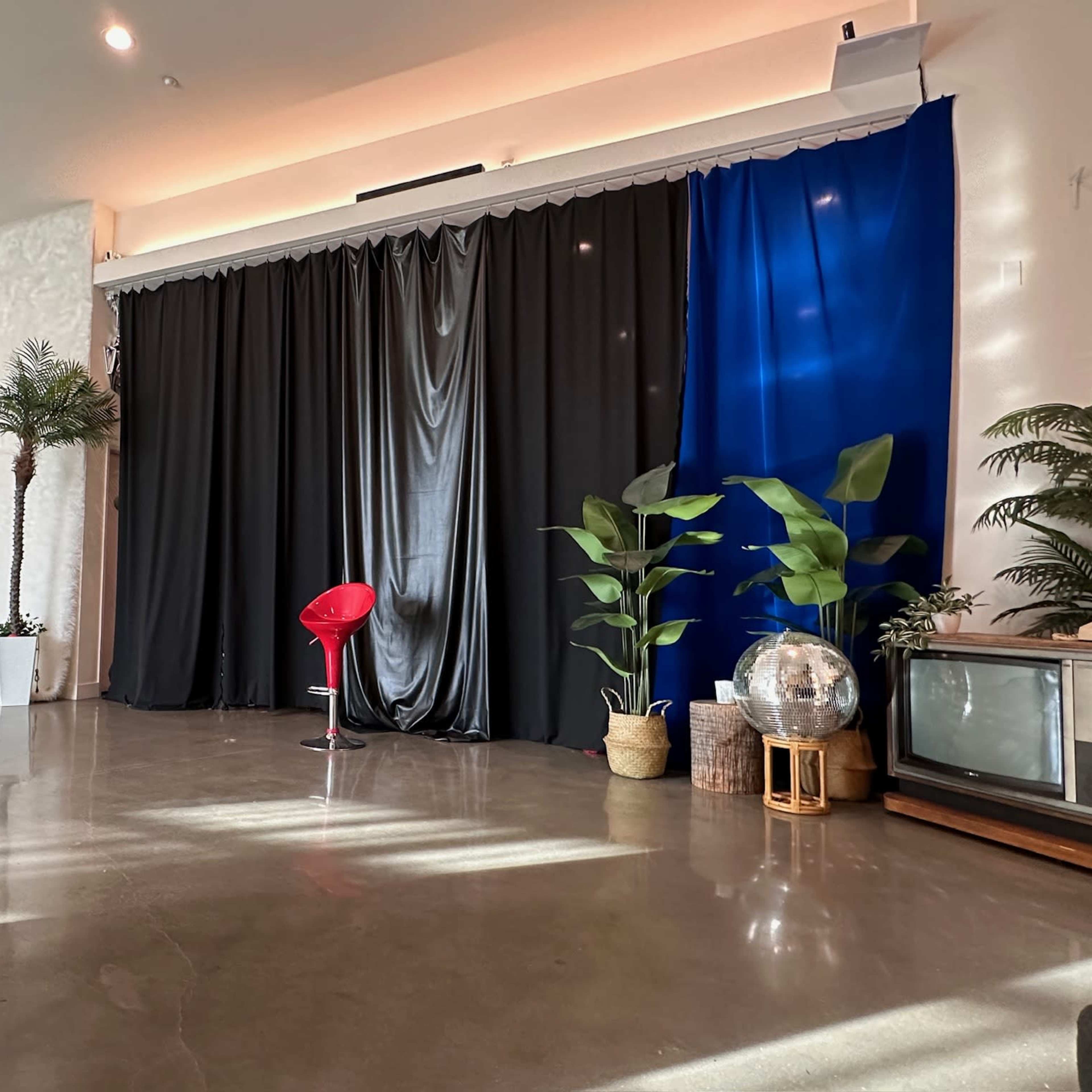 The scene features a modern interior space with a concrete floor, potted plants, a red chair, and a mix of black and blue curtains as a backdrop.