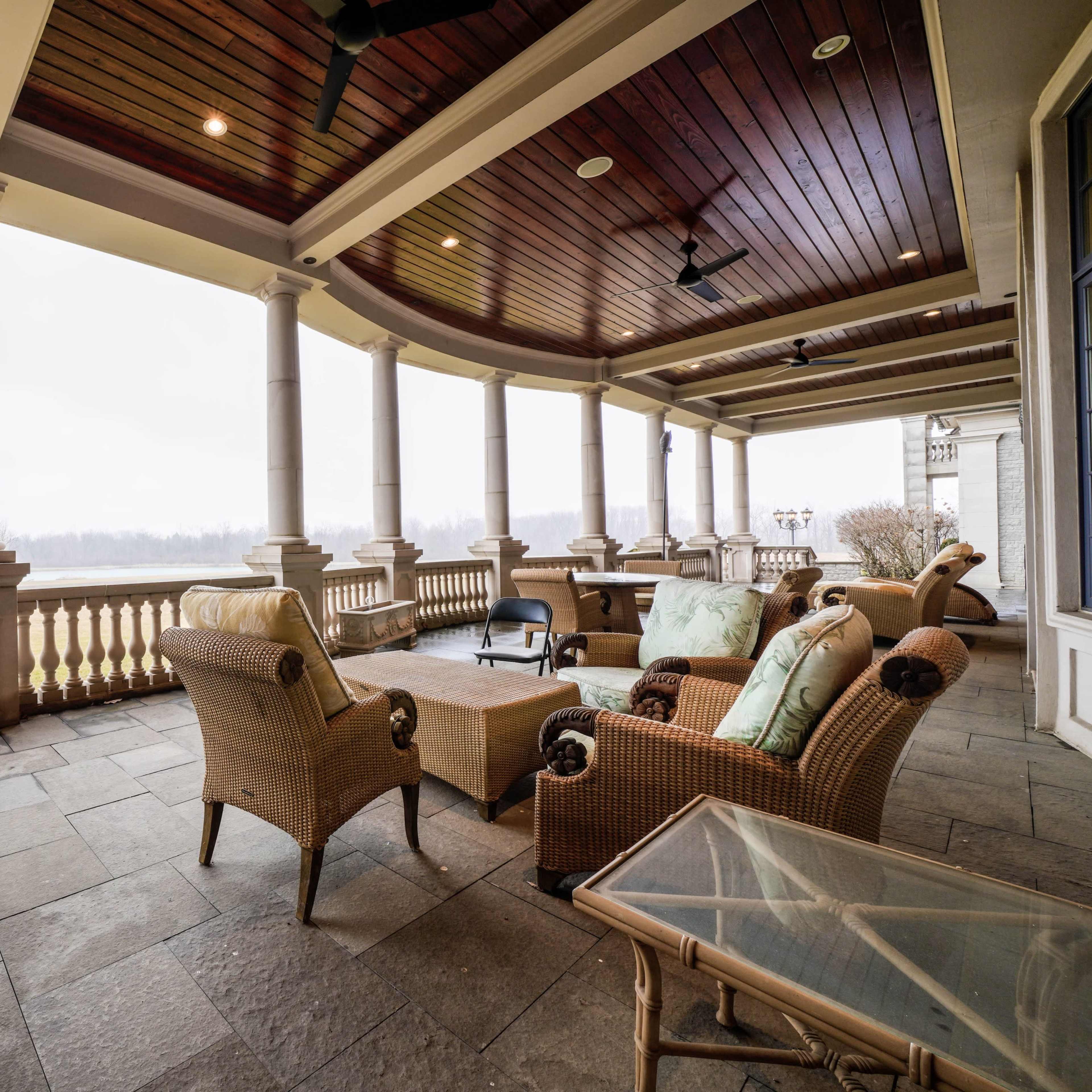 A covered porch with stone flooring, featuring wicker chairs and a glass-top table, overlooking a grassy landscape in the background.