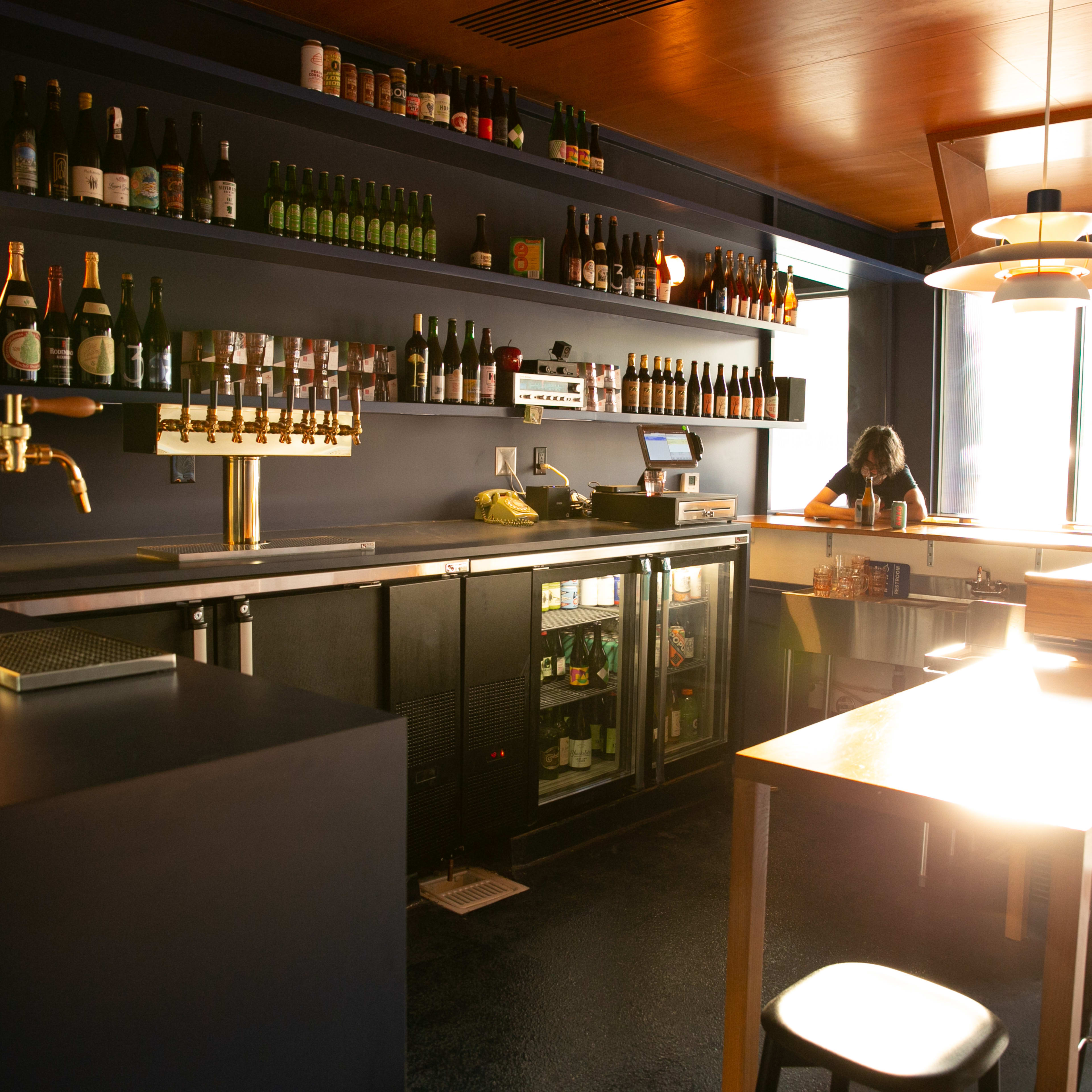 A modern bar area with a counter, shelves stocked with bottled drinks, and a bartender preparing in the background.