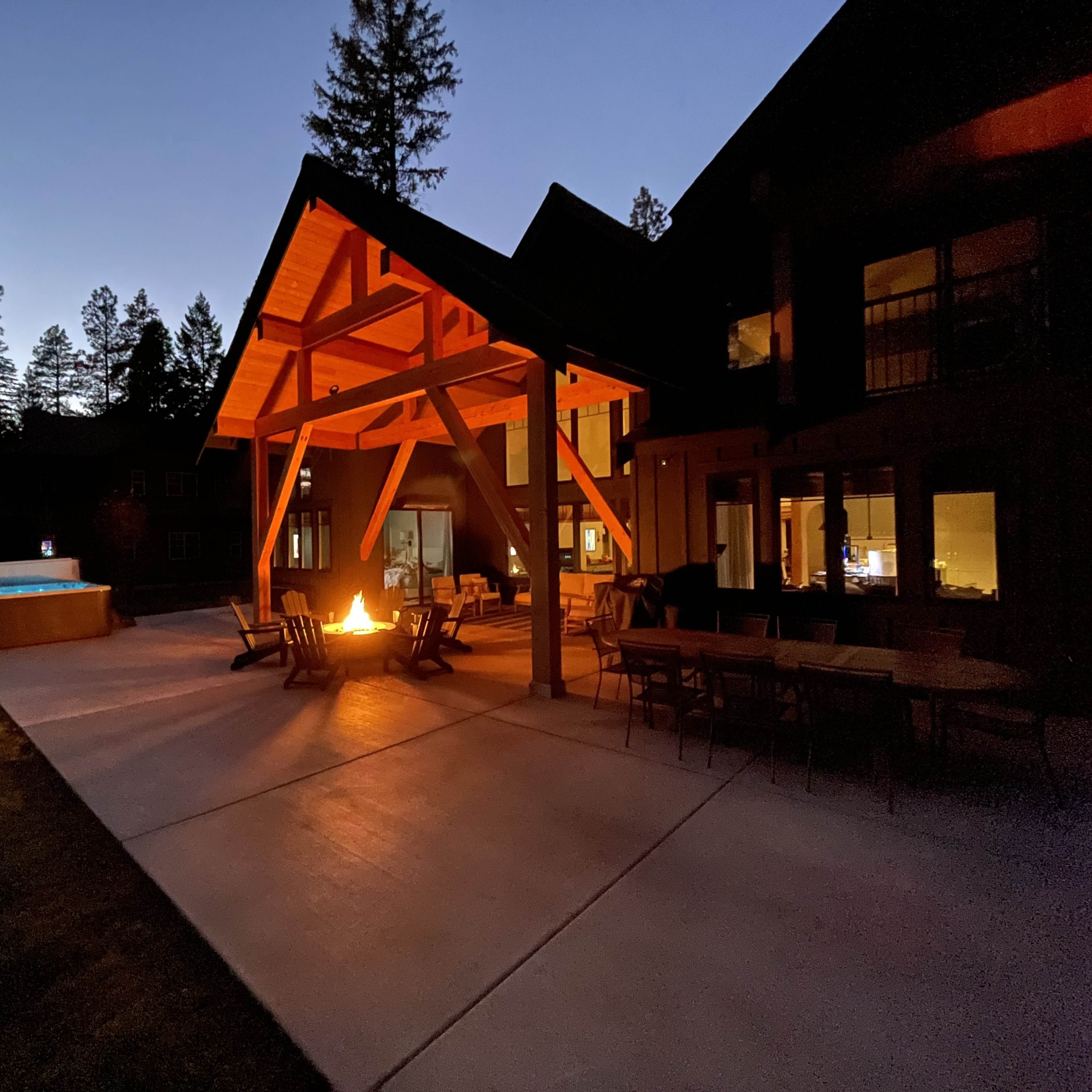 A modern outdoor patio features a fire pit surrounded by chairs, with a hot tub nearby, all illuminated by the warm glow of the house against a twilight sky.