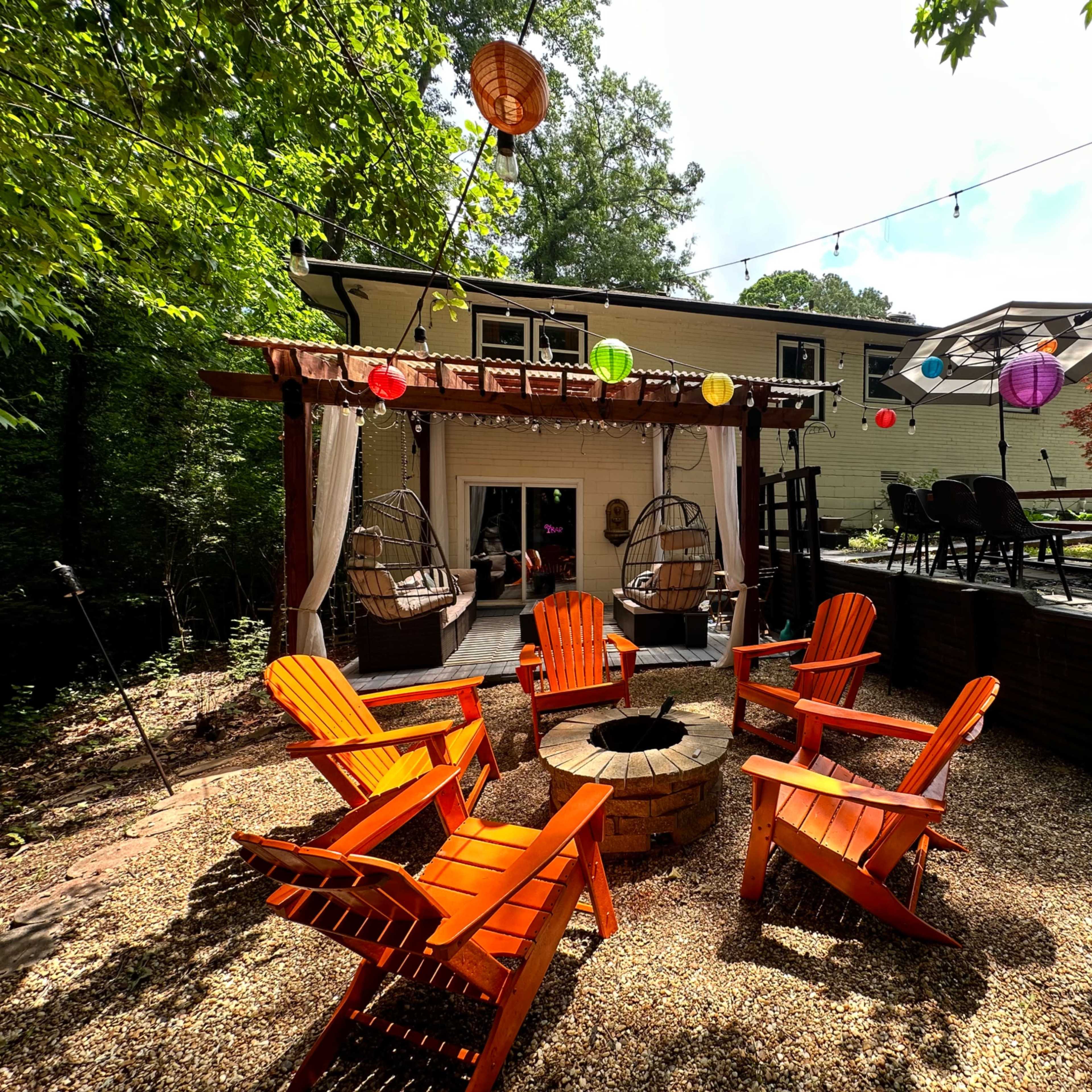 The scene shows a backyard with a stone fire pit surrounded by orange Adirondack chairs, hanging lanterns, and a patio area with a dining table.